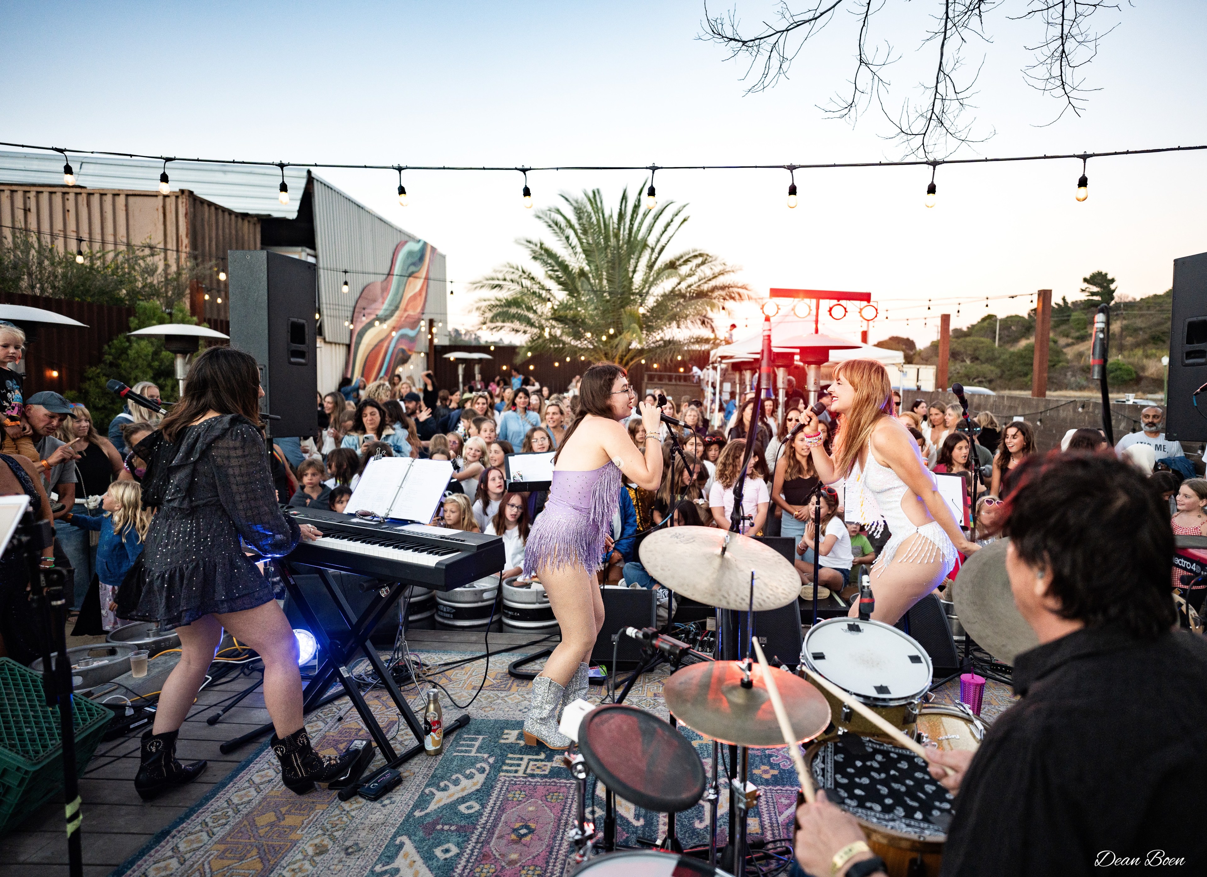 Musicians perform energetically on an outdoor stage at sunset, with a lively crowd gathered around. Strings of lights hang overhead, and palm trees are visible.