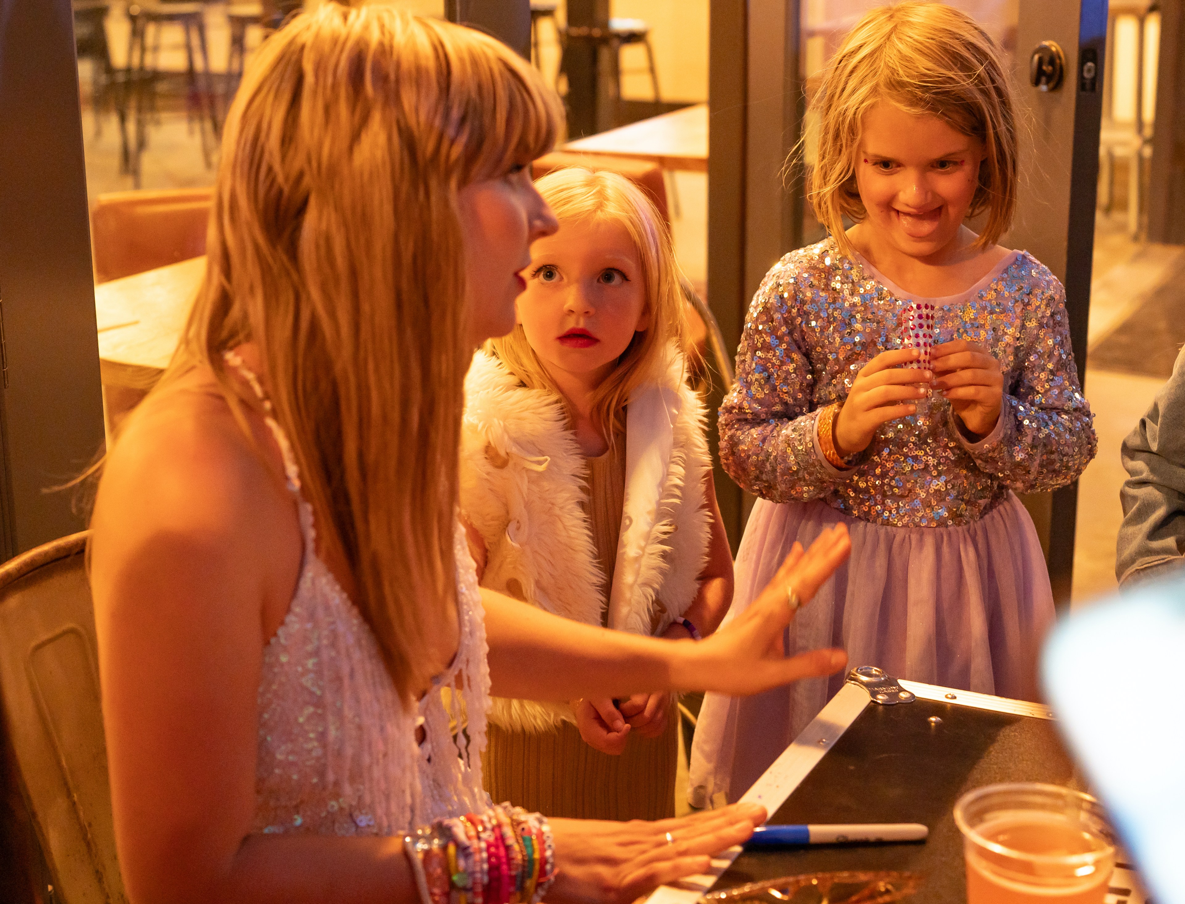 A woman in glittery attire talks to two children dressed in sparkly outfits. One child holds craft supplies while they appear engaged in a creative activity.