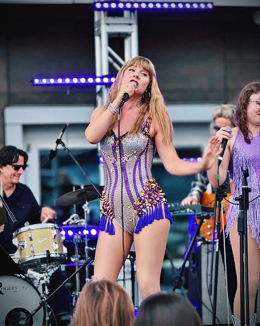 A singer in a sparkly purple and silver outfit performs on stage with a microphone, surrounded by other musicians and instruments.