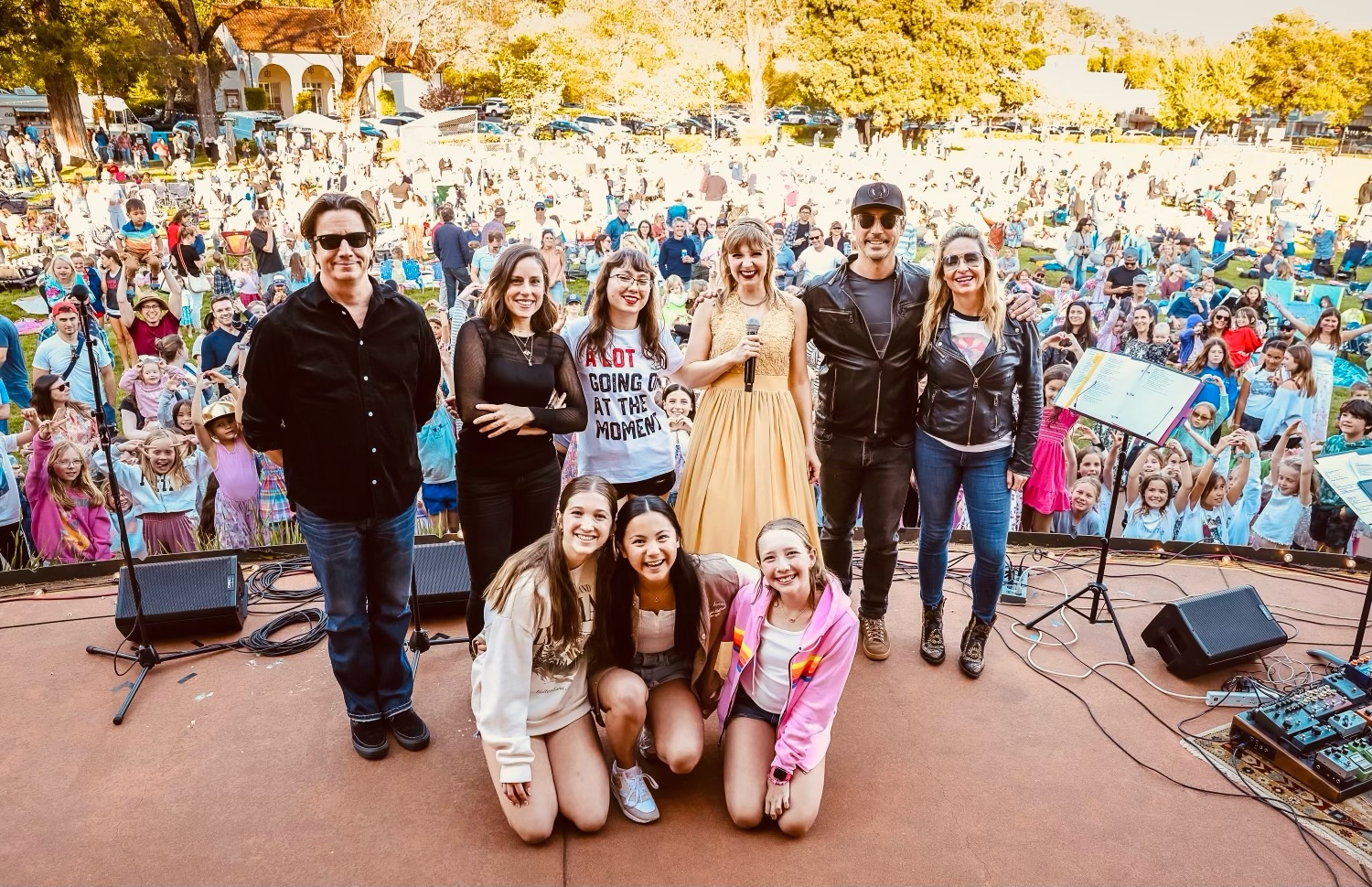 A group of nine people stand on a stage facing the camera, smiling with a large, lively crowd in the background. The setting is outdoors with trees and sunlight.