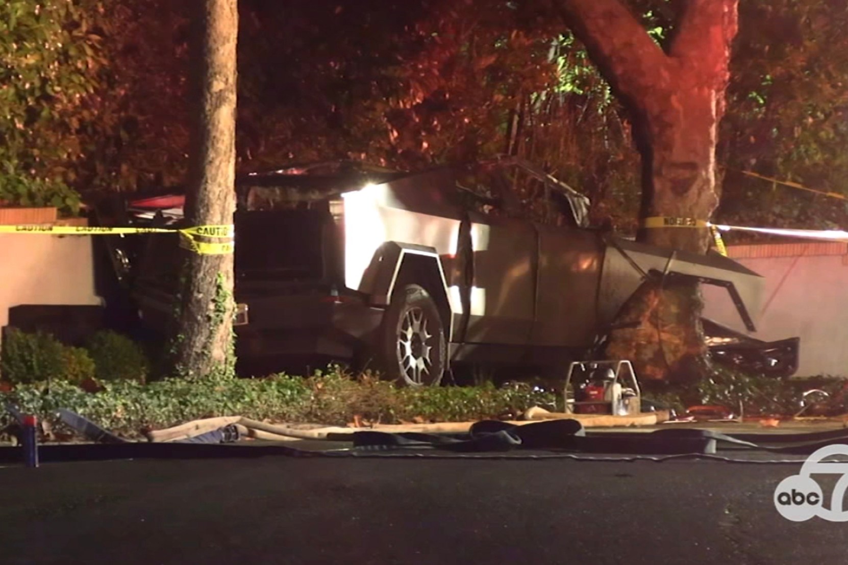 A damaged car is crashed into a tree beside a wall, surrounded by caution tape. It appears to be nighttime, with some debris on the ground.