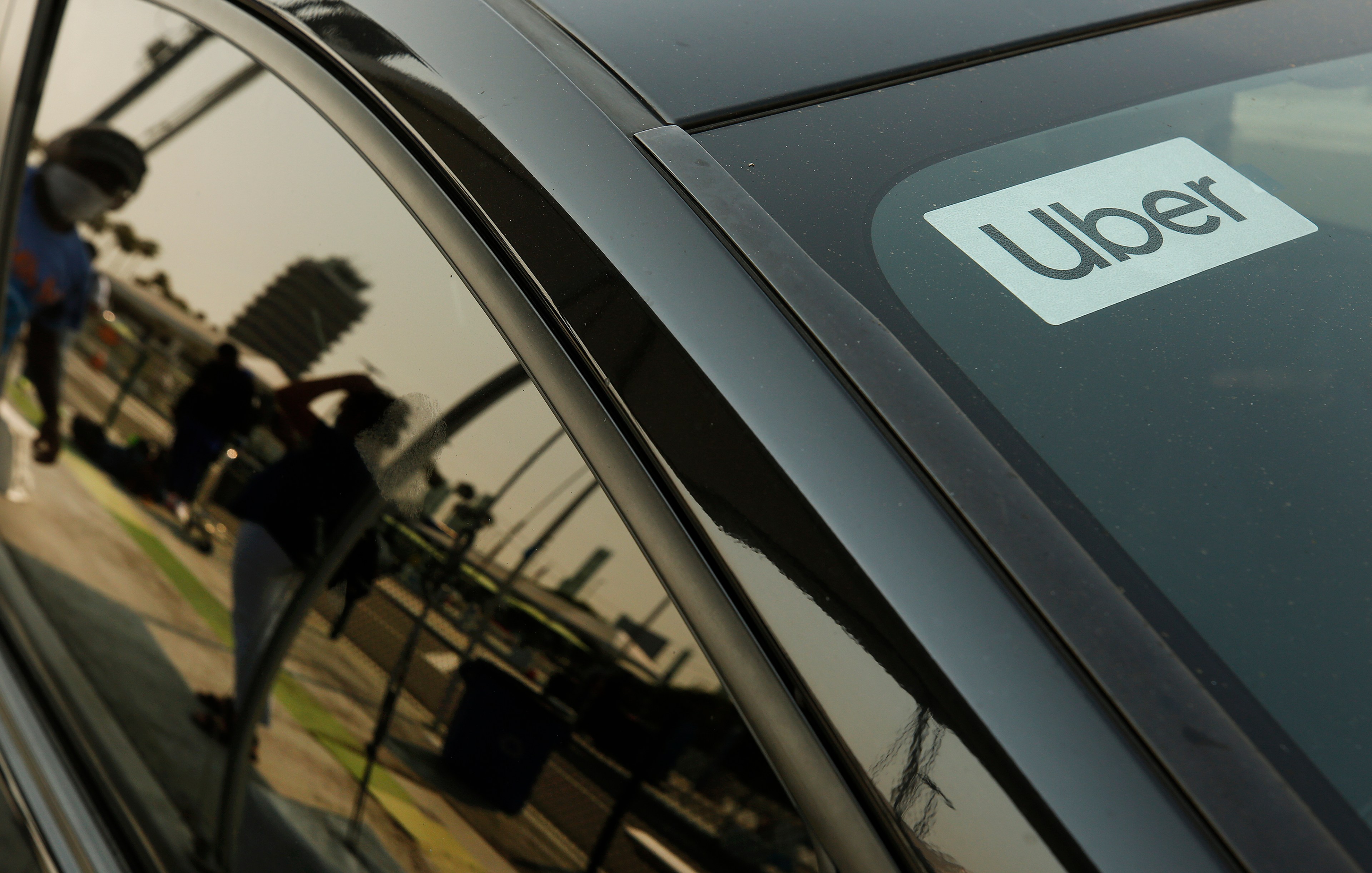 A black car displays an Uber sign in the window. The reflection on the car shows blurred people and a building under a sunny sky.