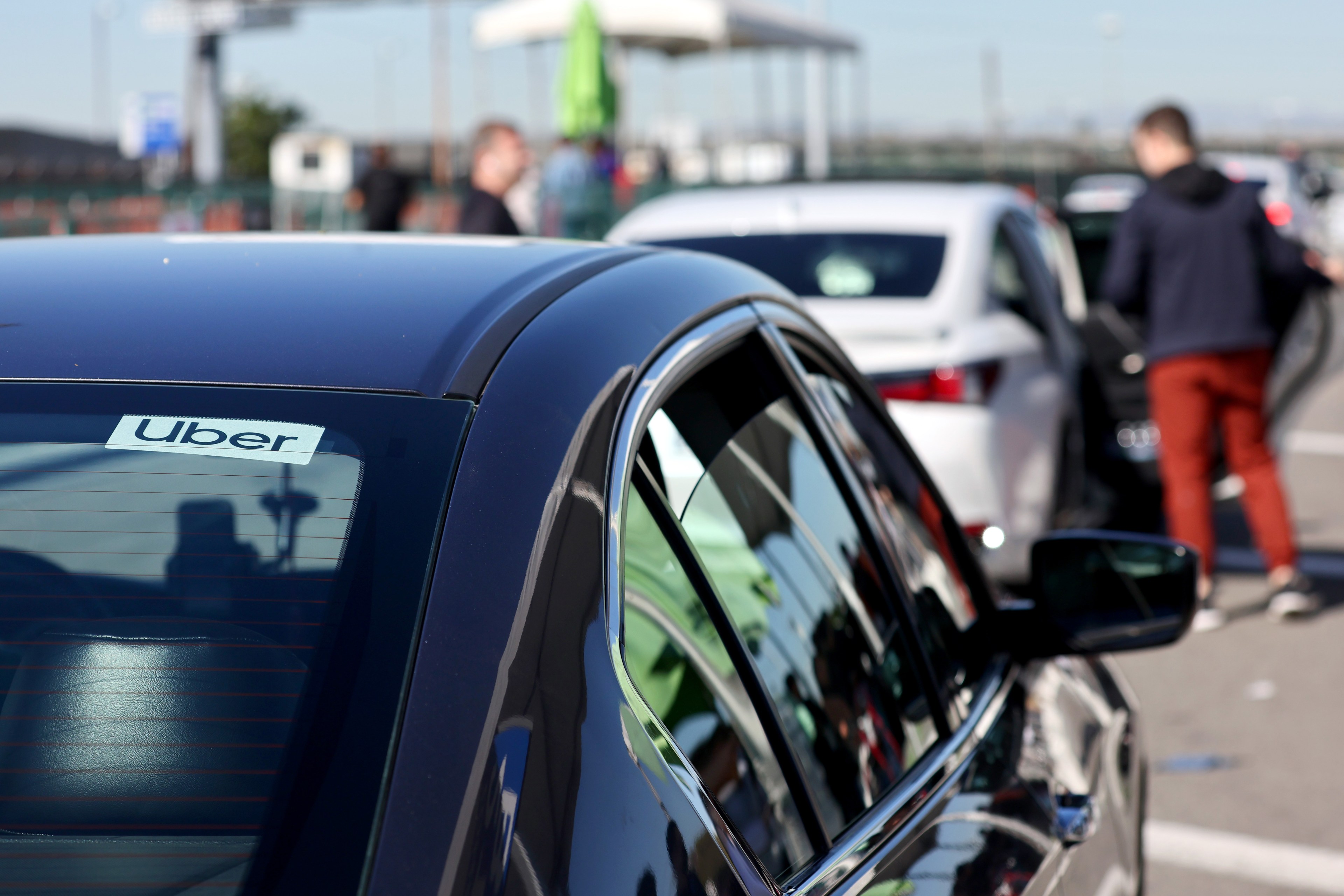 A parked black car with an Uber sticker on the rear window is in focus, while people and other vehicles are blurred in the background on a sunny day.