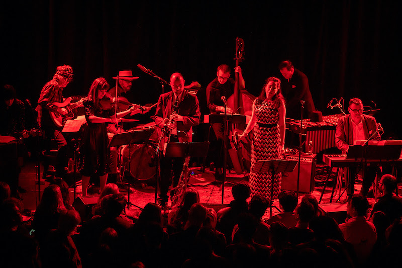 A band performs on stage under red lighting, featuring musicians playing saxophone, guitar, violin, bass, and keyboards, with a vocalist and visible audience.