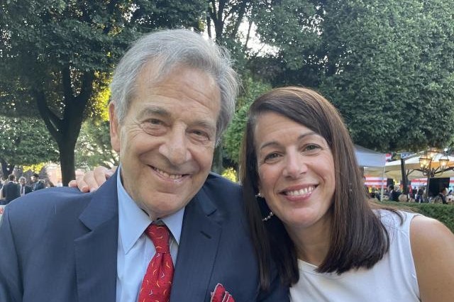 A man in a dark suit with a red tie and pocket square smiles alongside a woman in a white dress. They are outdoors with trees and people in the background.