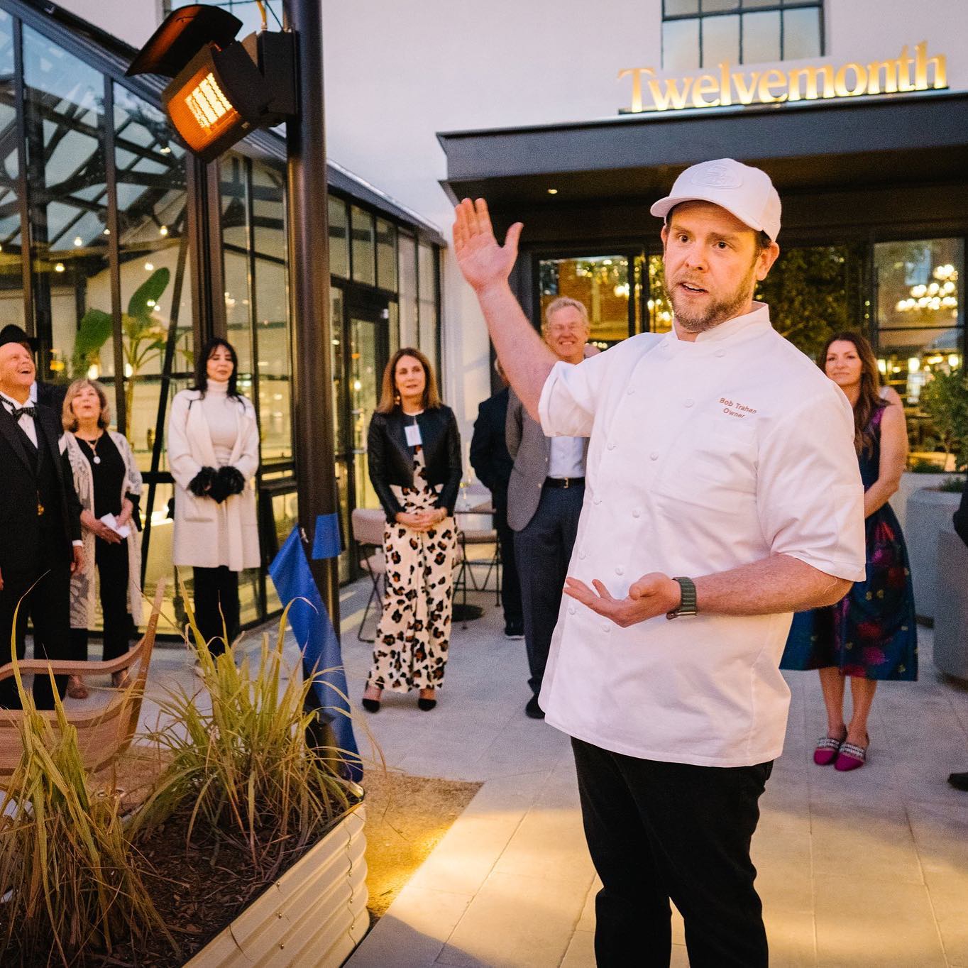 A chef in a white uniform gestures animatedly outdoors, surrounded by a group of well-dressed people near a restaurant with &quot;Twelvemonth&quot; on the sign.