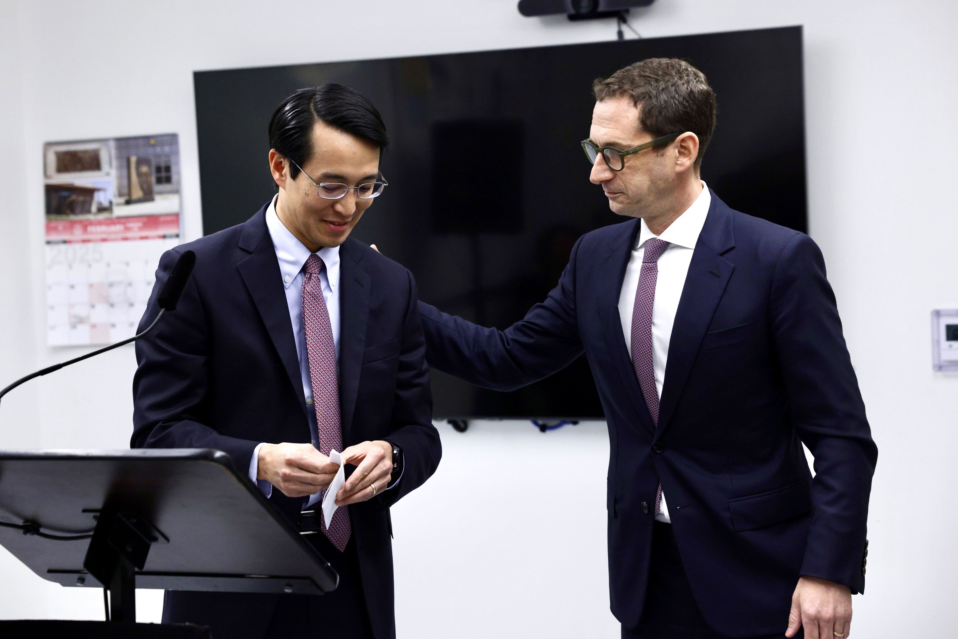 Two men in suits stand near a podium. One is holding papers, and the other rests a hand on his shoulder. A calendar and TV are visible in the background.