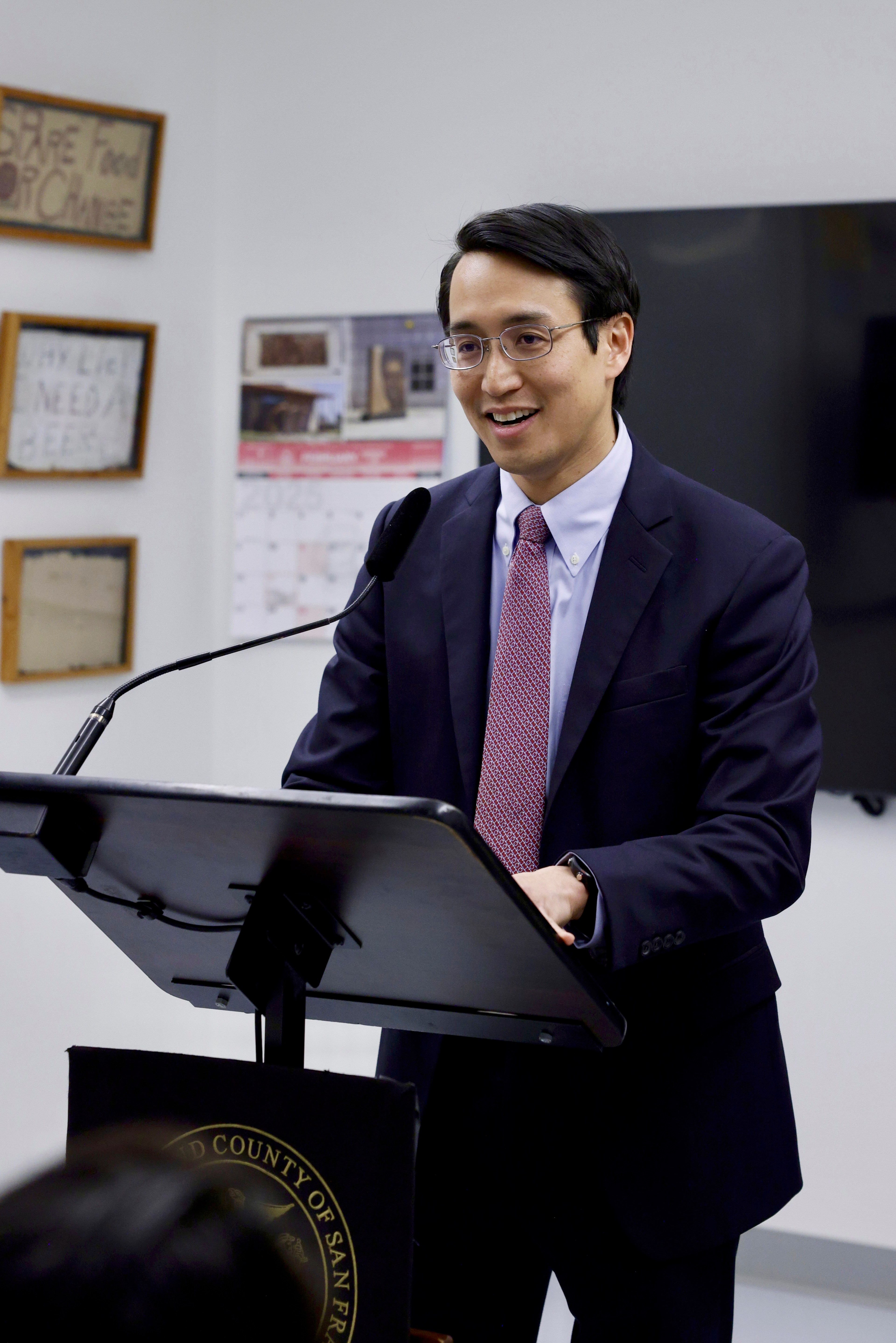 A man in a suit and tie is speaking at a podium with a microphone. There's a calendar and framed signs on the wall behind him.