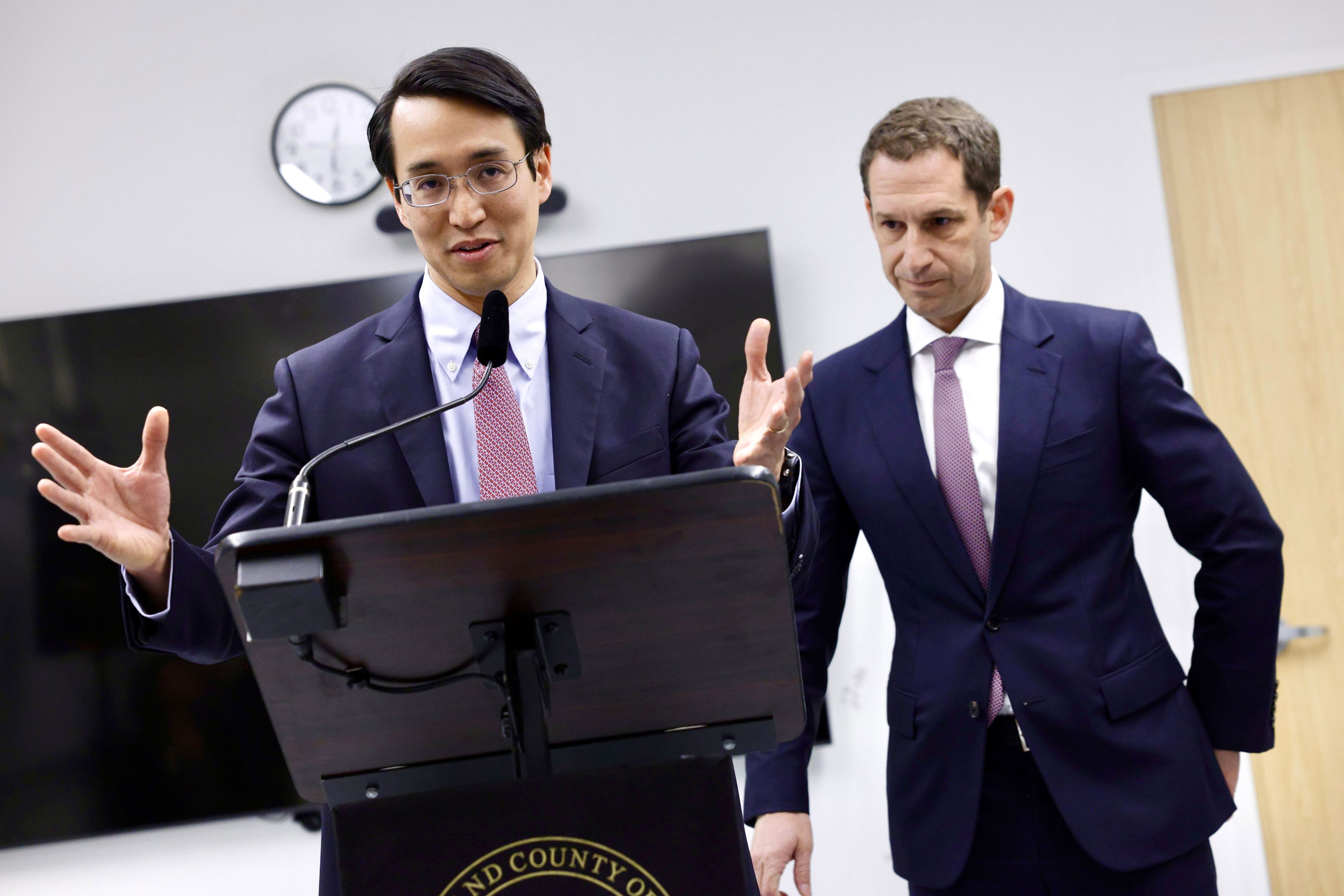 A person in a suit speaks at a podium with a microphone, gesturing with hands, while another person in a suit stands beside them, listening attentively.