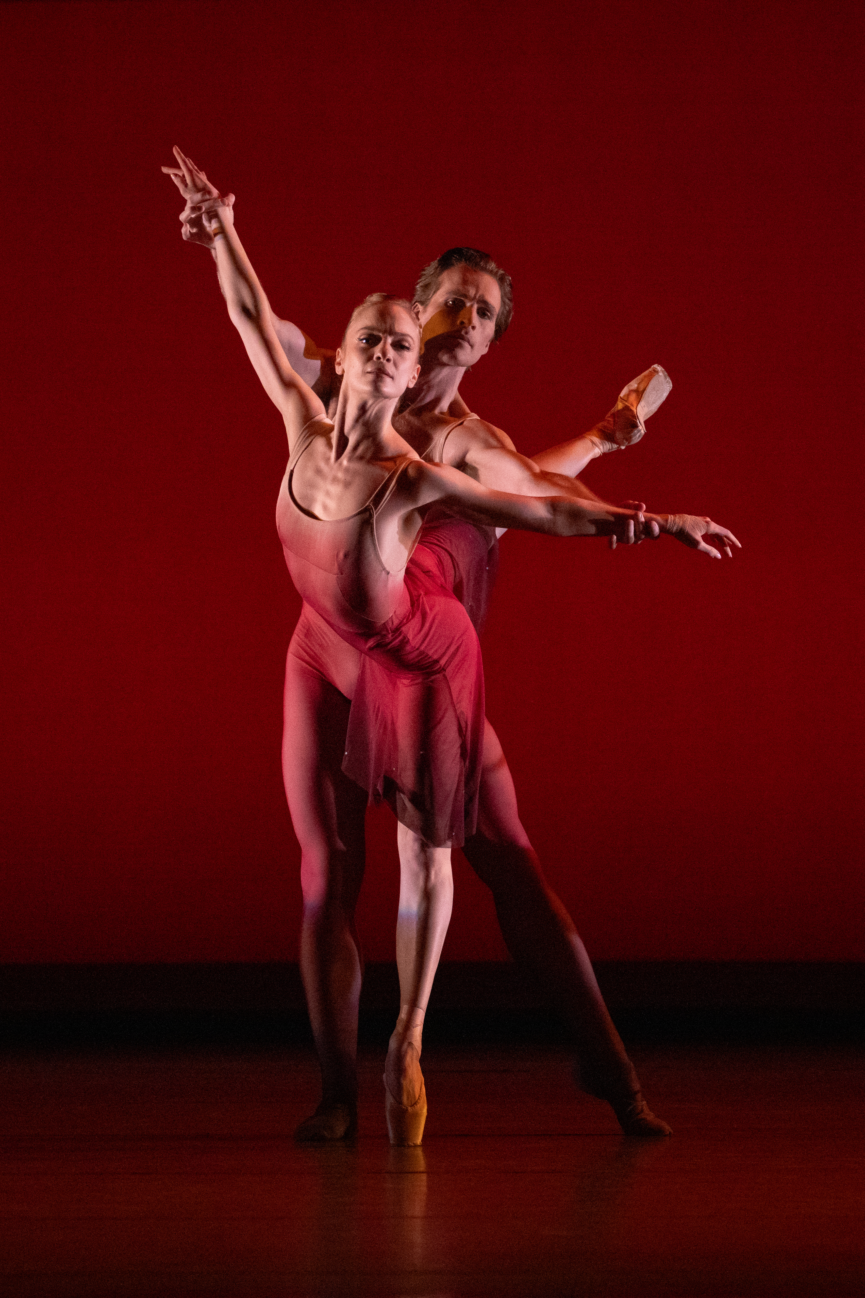 Two ballet dancers perform in front of a red backdrop. They wear matching red outfits, striking a graceful pose with extended arms and one standing en pointe.