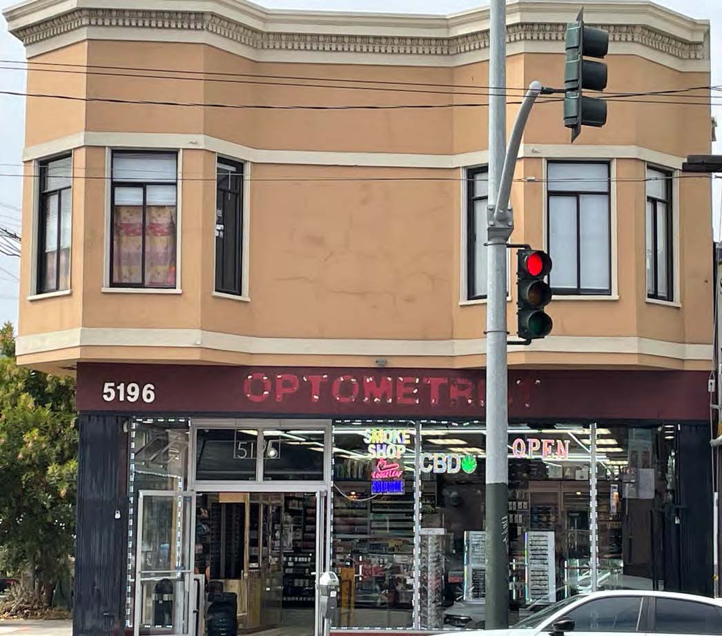 A two-story building with a corner shop labeled &quot;OPTOMETRY&quot; and &quot;Smoke Shop.&quot; The storefront is open, displaying neon signs, with a traffic light nearby.