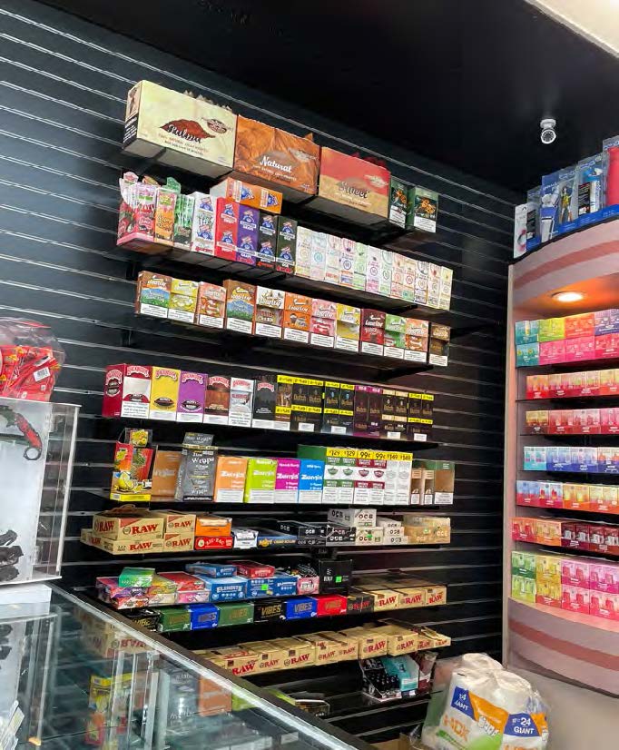 The image shows a store display with shelves filled with various brands of rolling papers and smoking accessories. The background is black, and items are neatly arranged.