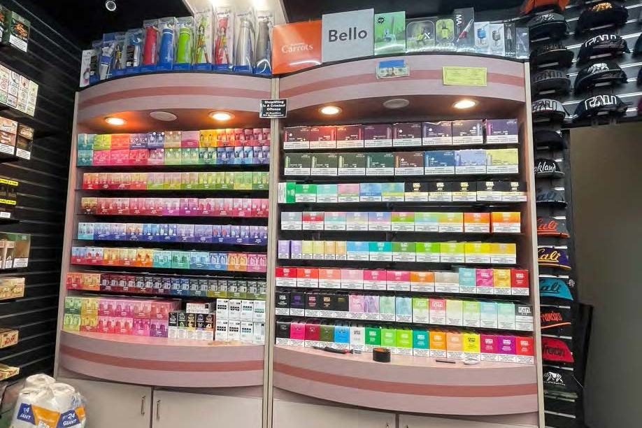 The image shows a store display with shelves full of colorful vape or e-cigarette products and accessories beneath bright lights. Hats are displayed on the right.