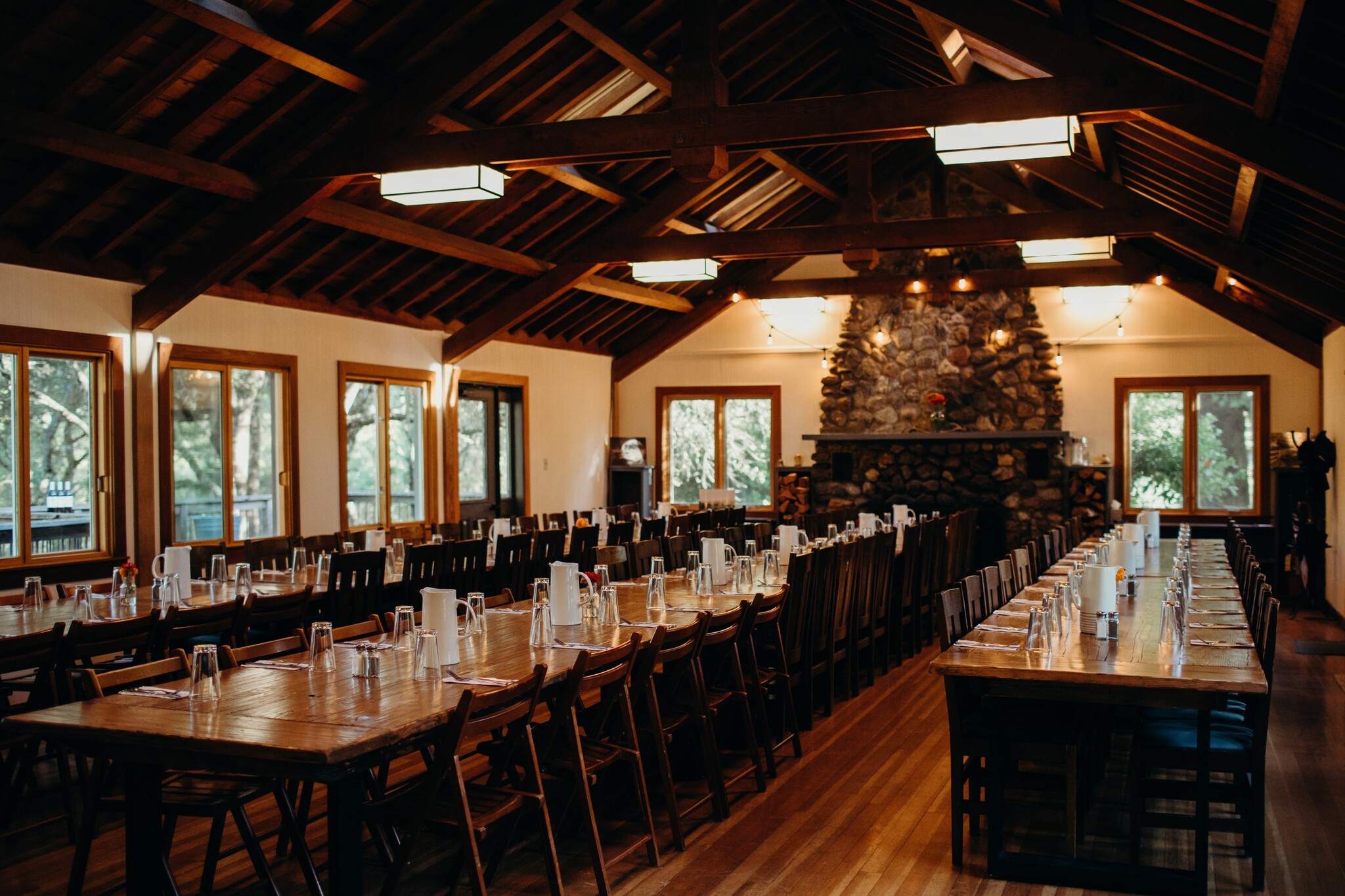 The image shows a large, rustic dining hall with wooden beams and tables set for a meal. There's a stone fireplace at the end, and windows line the walls.