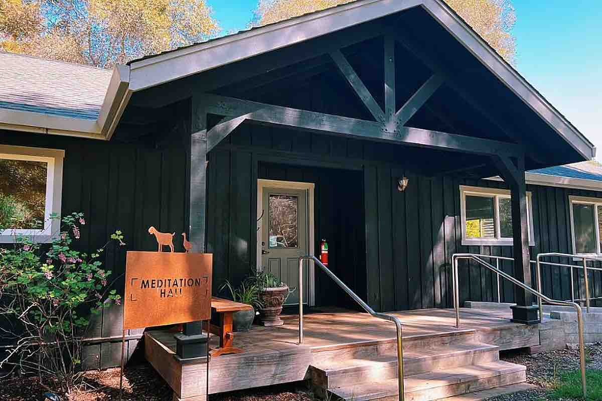 A wooden building, labeled &quot;Meditation Hall,&quot; has a small porch with railings. There's a sign with a goat figure, surrounded by trees and plants.