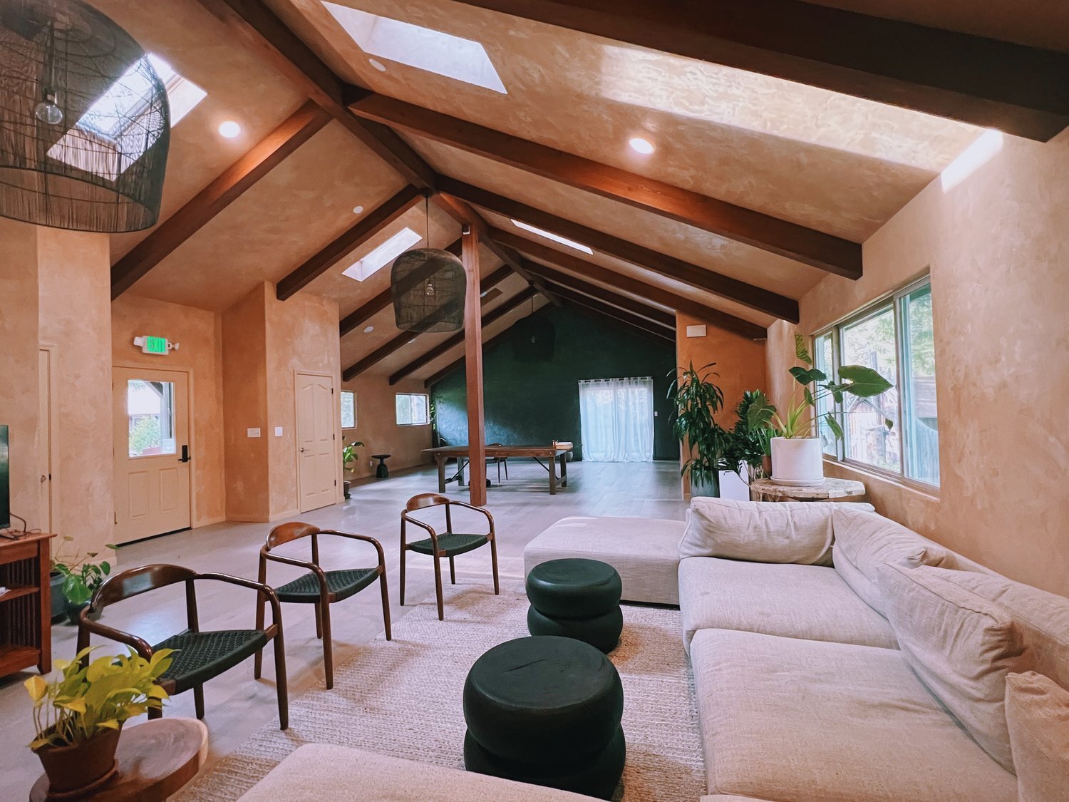 The image shows a spacious, modern living room with vaulted ceilings, skylights, a beige sectional sofa, wooden chairs, potted plants, and large windows.