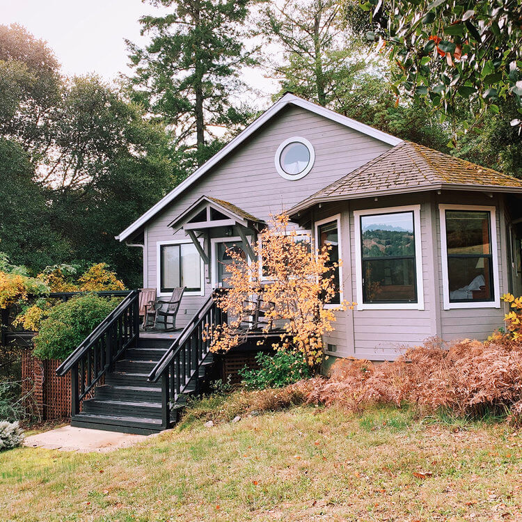 A small, charming house with gray siding stands amidst lush greenery. It features a porch with black railings, surrounded by trees and a grassy yard.