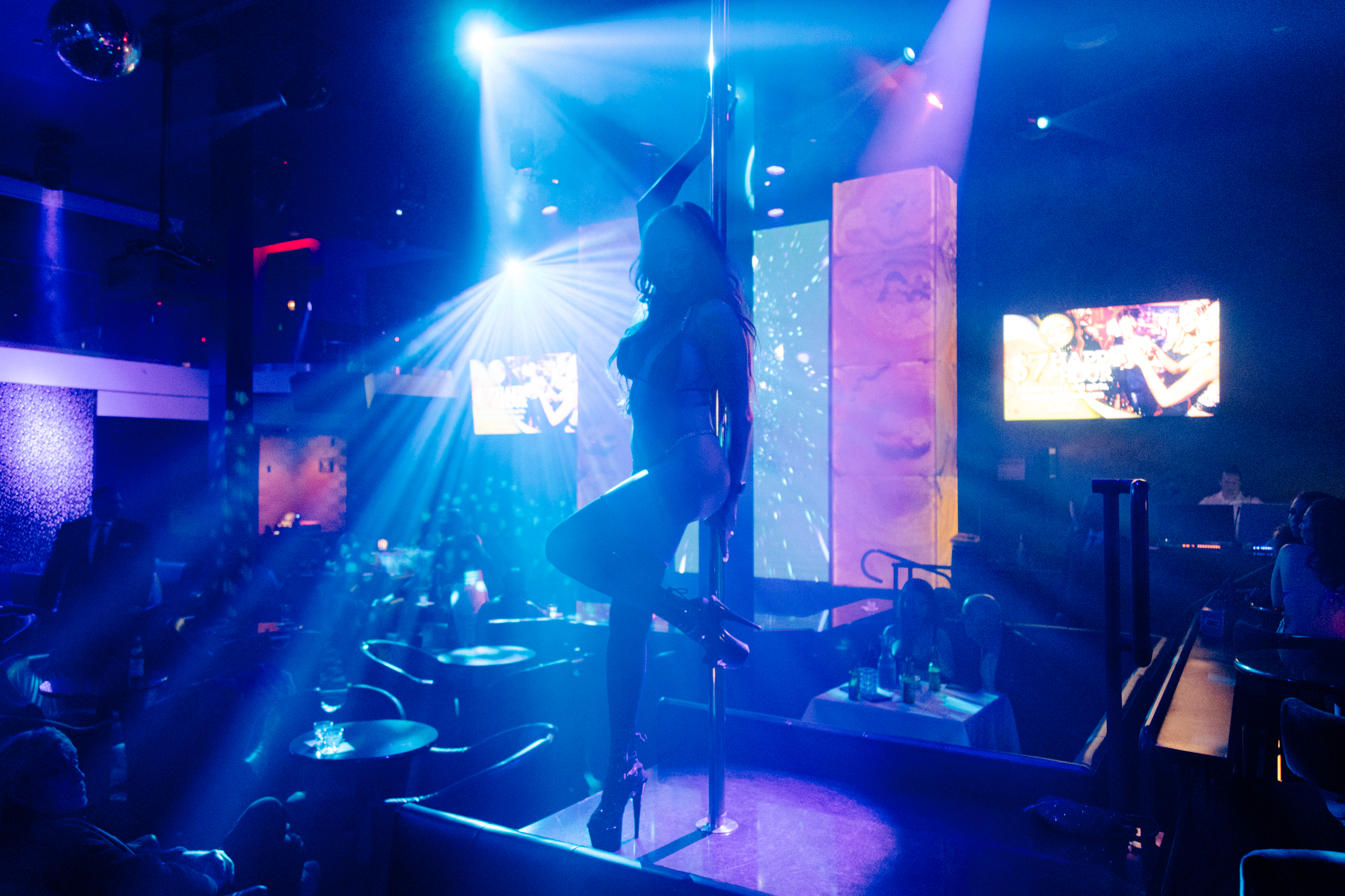 A dimly lit nightclub scene shows a performer dancing on a pole, illuminated by colorful lights. Patrons sit around tables, watching, with screens in the background.