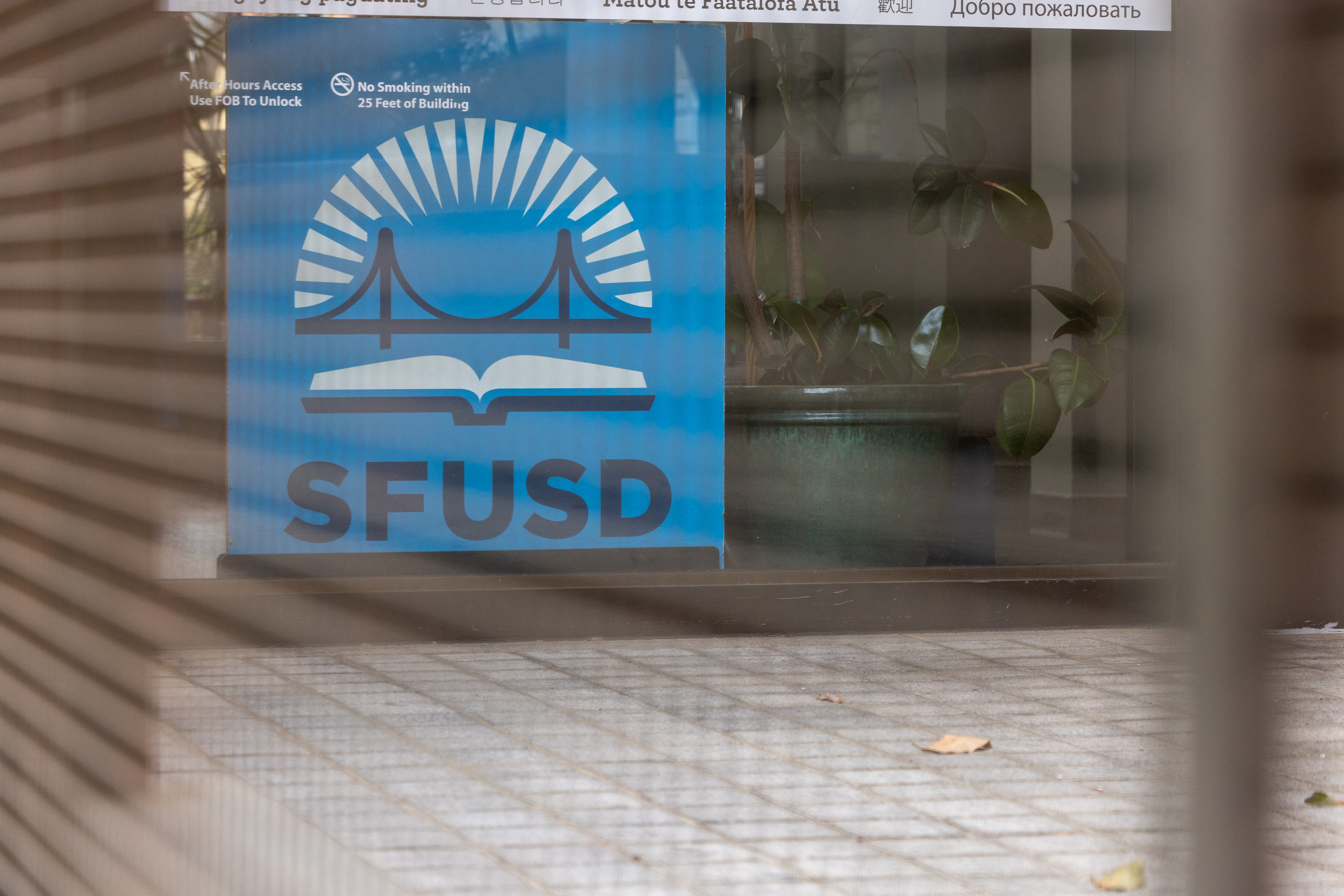 The image shows a sign with a blue background featuring a bridge and book logo, and the letters "SFUSD." It's viewed through slats with a plant nearby.