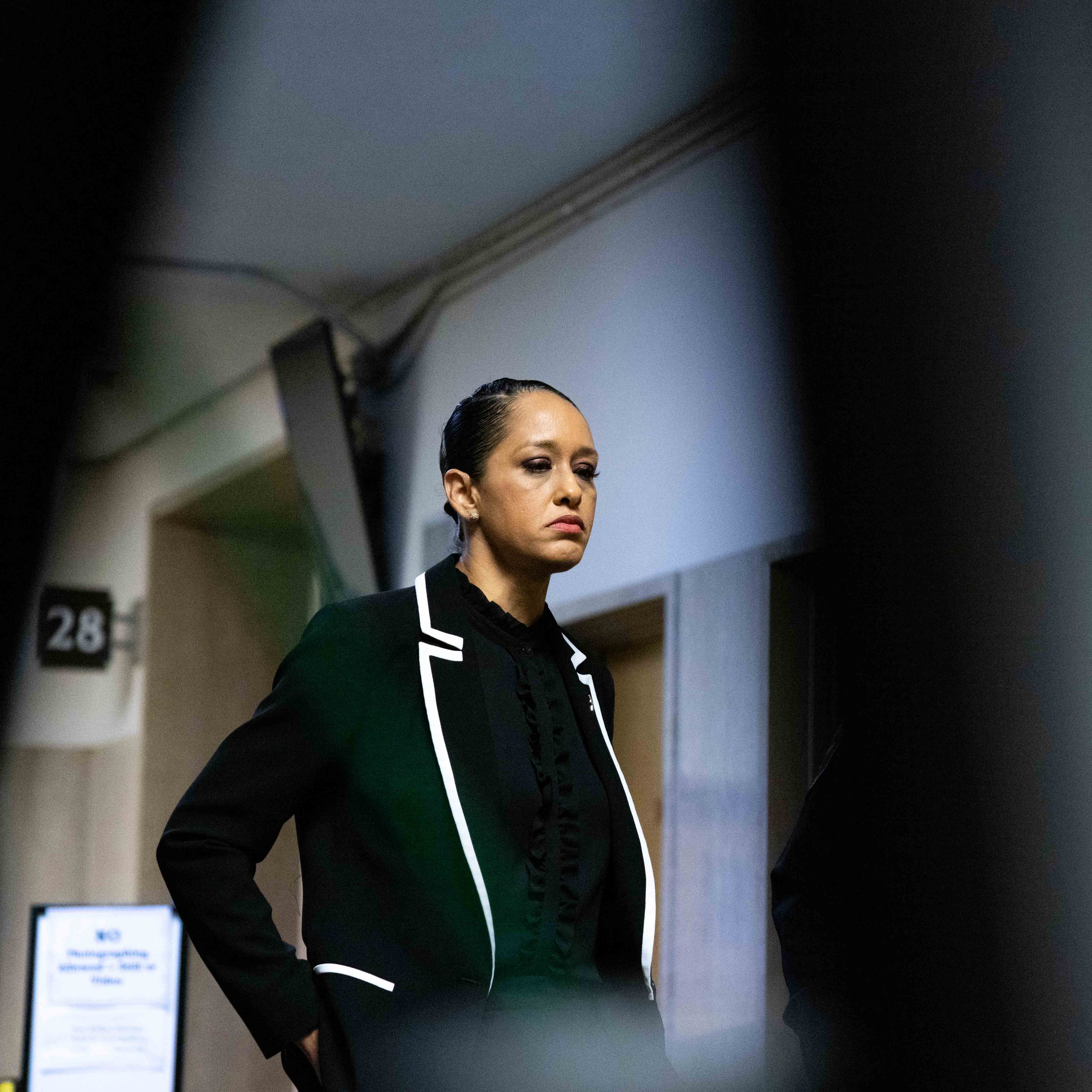 A woman in a black suit with white trim stands in a hallway, appearing serious. The image is framed by blurred foreground objects, creating a focused view.