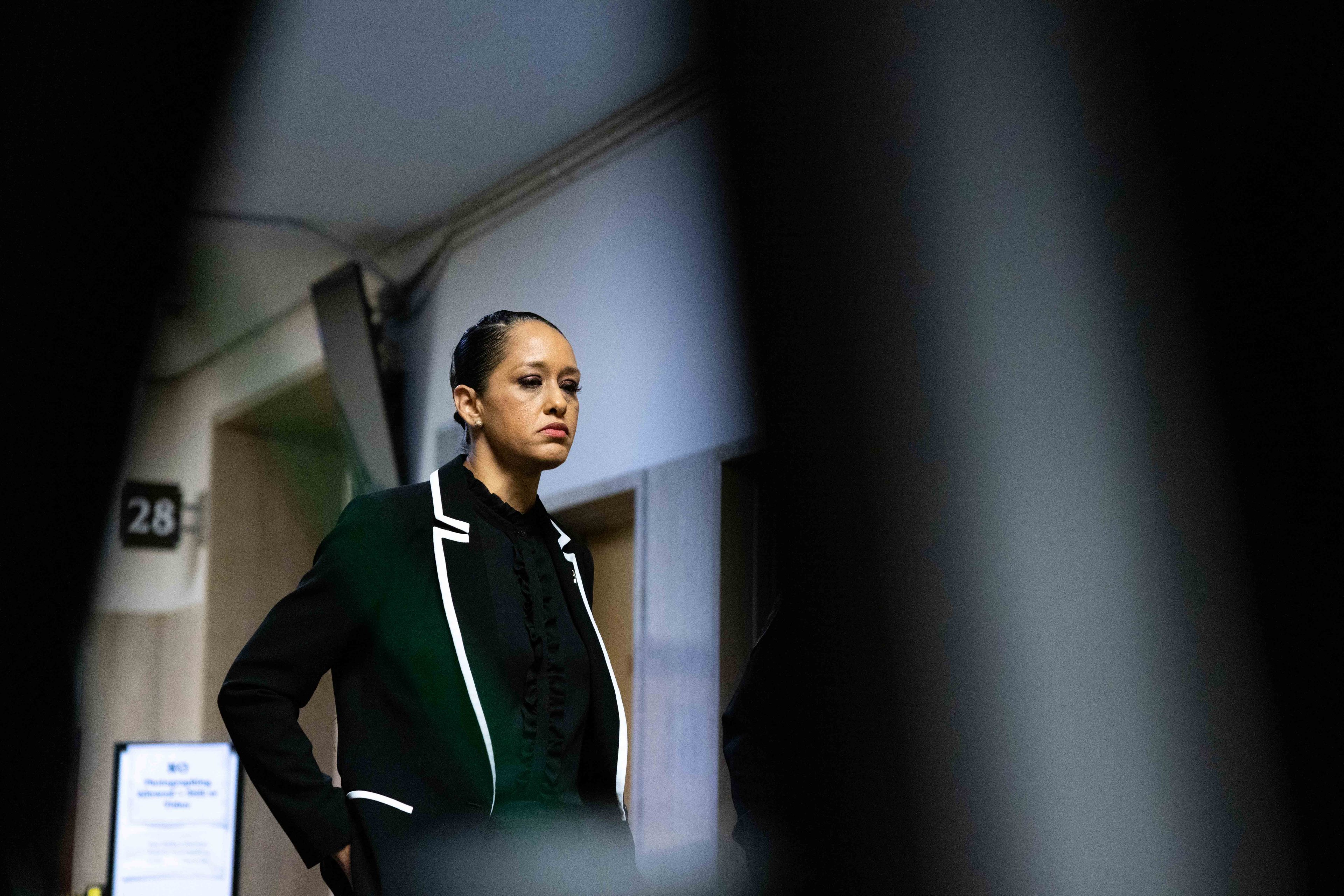 A woman in a black suit with white trim stands in a hallway, appearing serious. The image is framed by blurred foreground objects, creating a focused view.