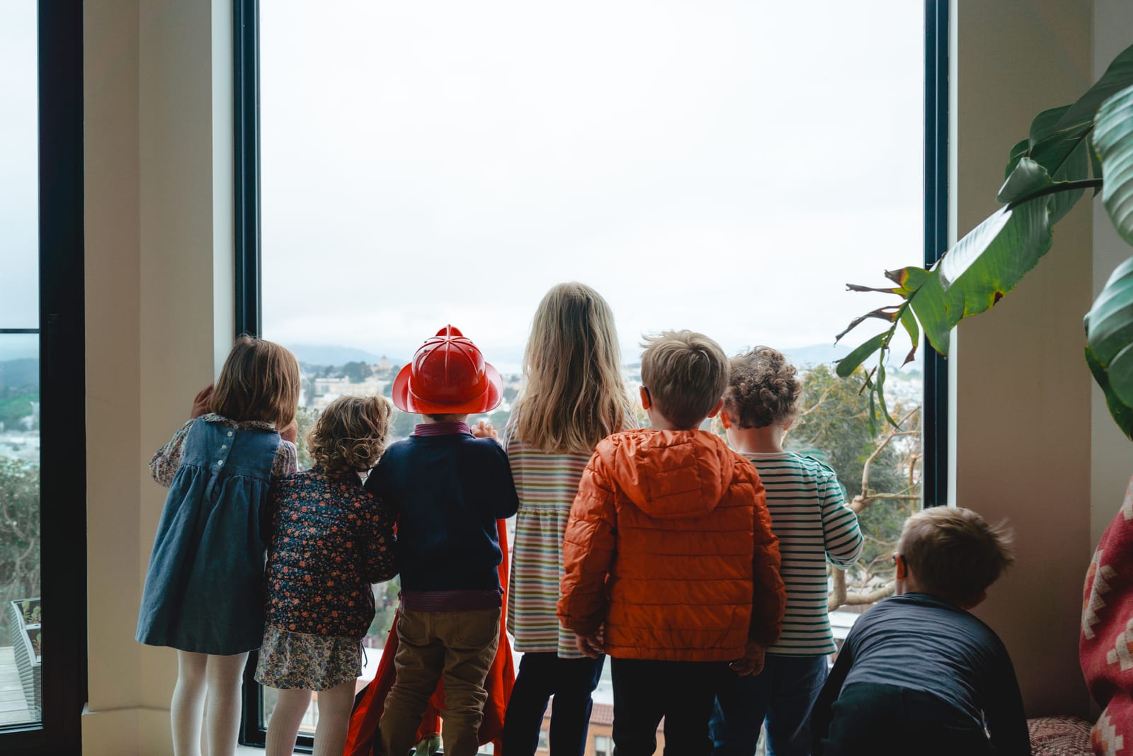 Seven children, with backs turned, stand by a large window, gazing outside. One wears a red firefighter helmet. Indoor plants are visible beside them.