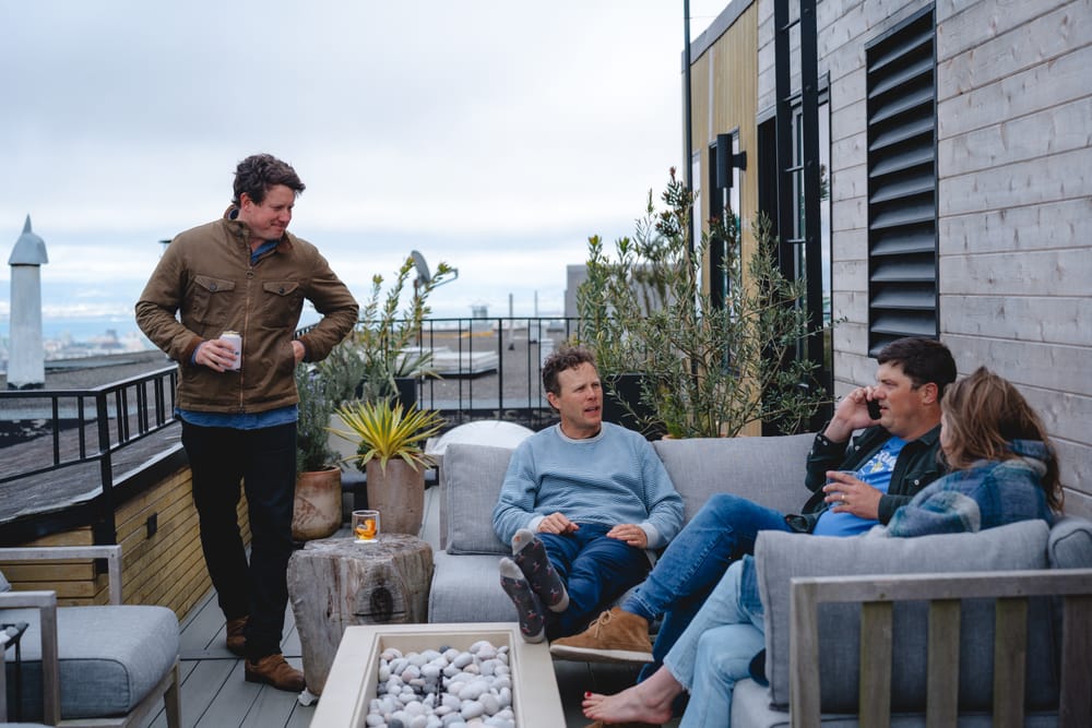 Four people are on a rooftop patio. Three sit on a sofa, with one on a phone, and one stands holding a drink. The setting is casual with plants and a fire pit.