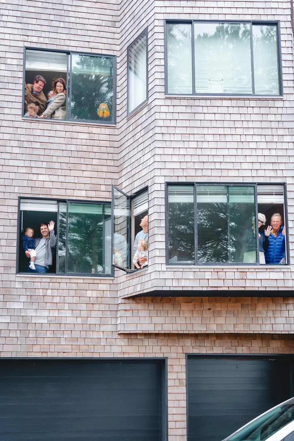 People of various ages are looking out from multiple windows of a wooden house's upper floor, with some waving and smiling. There's a glimpse of garage doors below.