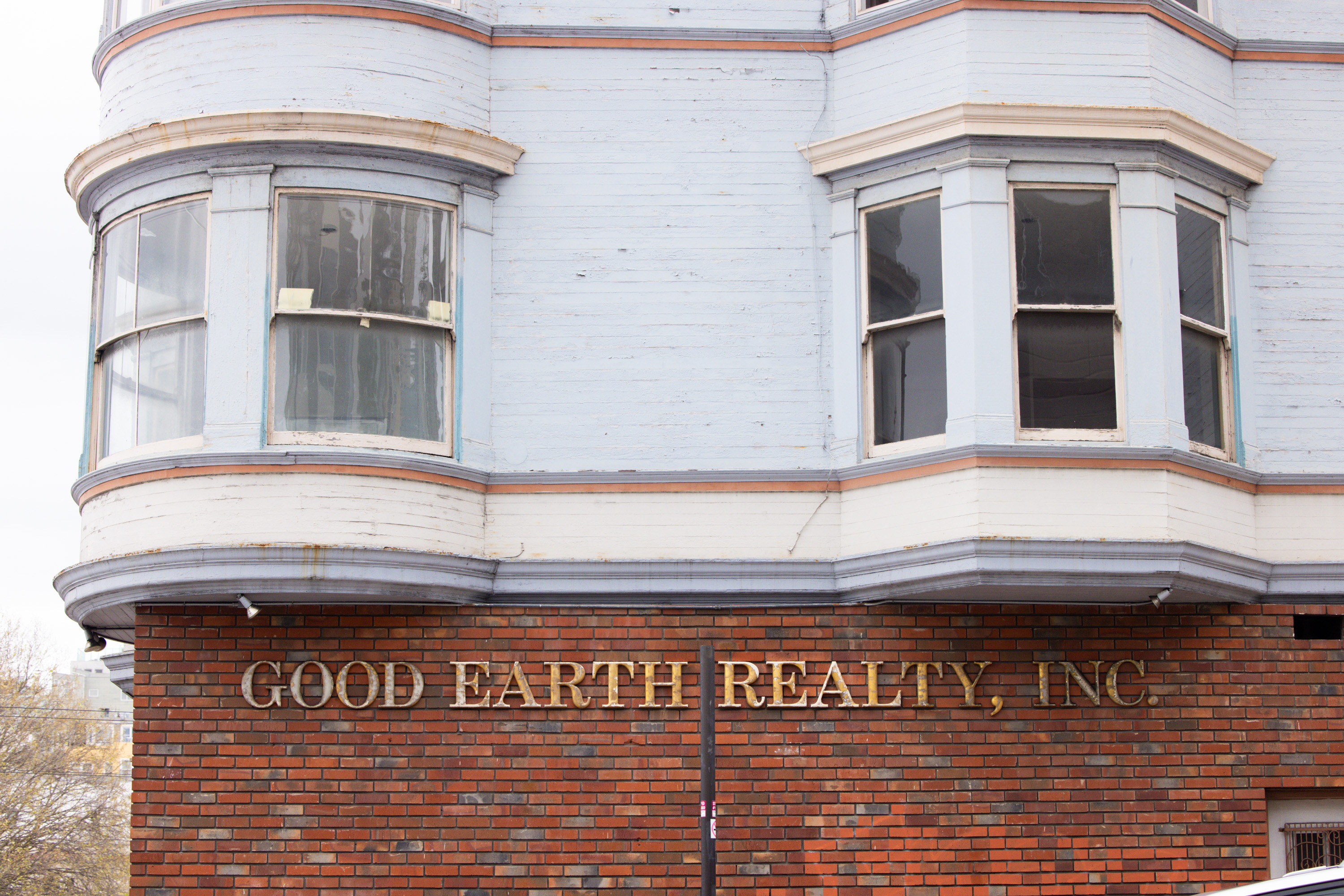 A building features bay windows with light blue wooden siding. Below, red brick walls display the gold-lettered sign &quot;Good Earth Realty, Inc.&quot;