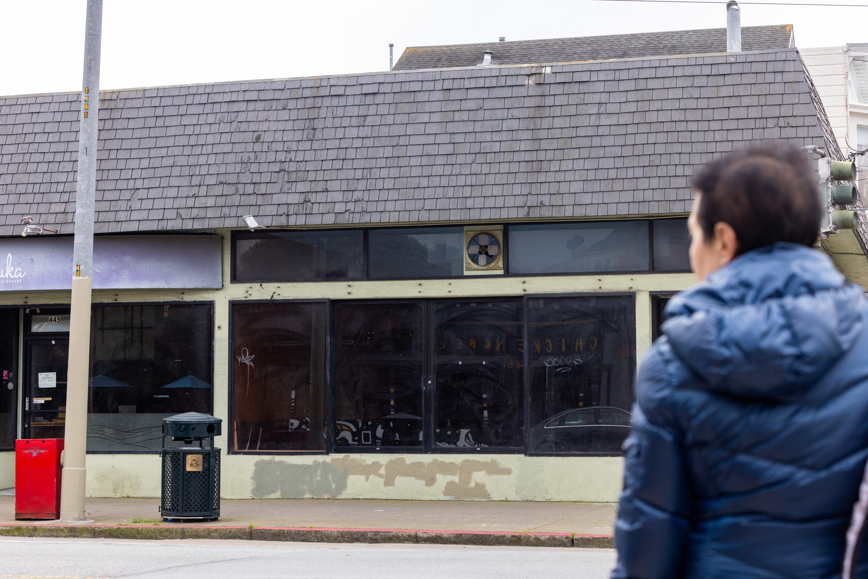 A person in a blue jacket stands on a street in front of a building with large windows, a shingled roof, and faded signage. An empty trash can is nearby.