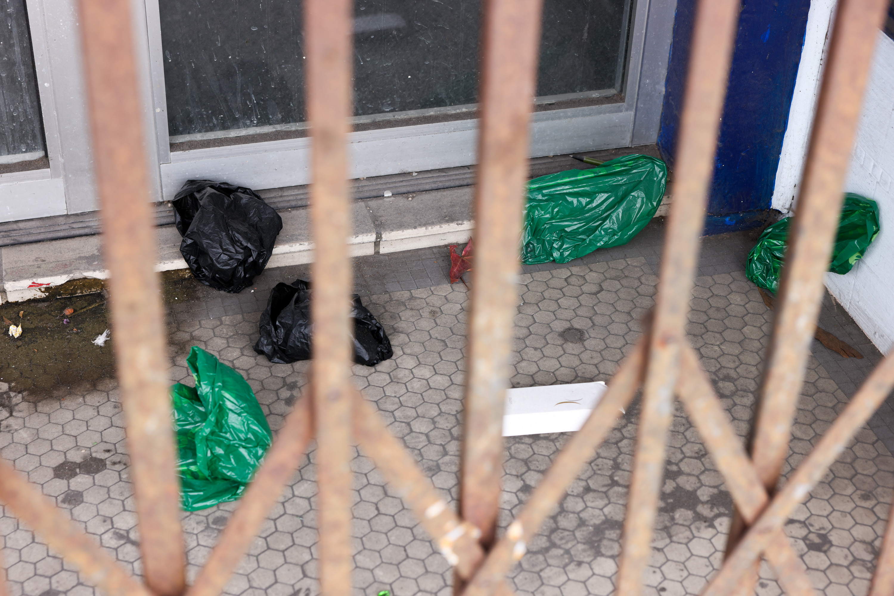 A barred gate with sacks of green and black plastic bags is set on a grimy hexagonal-tiled floor. A white rectangular object lies near the bags.