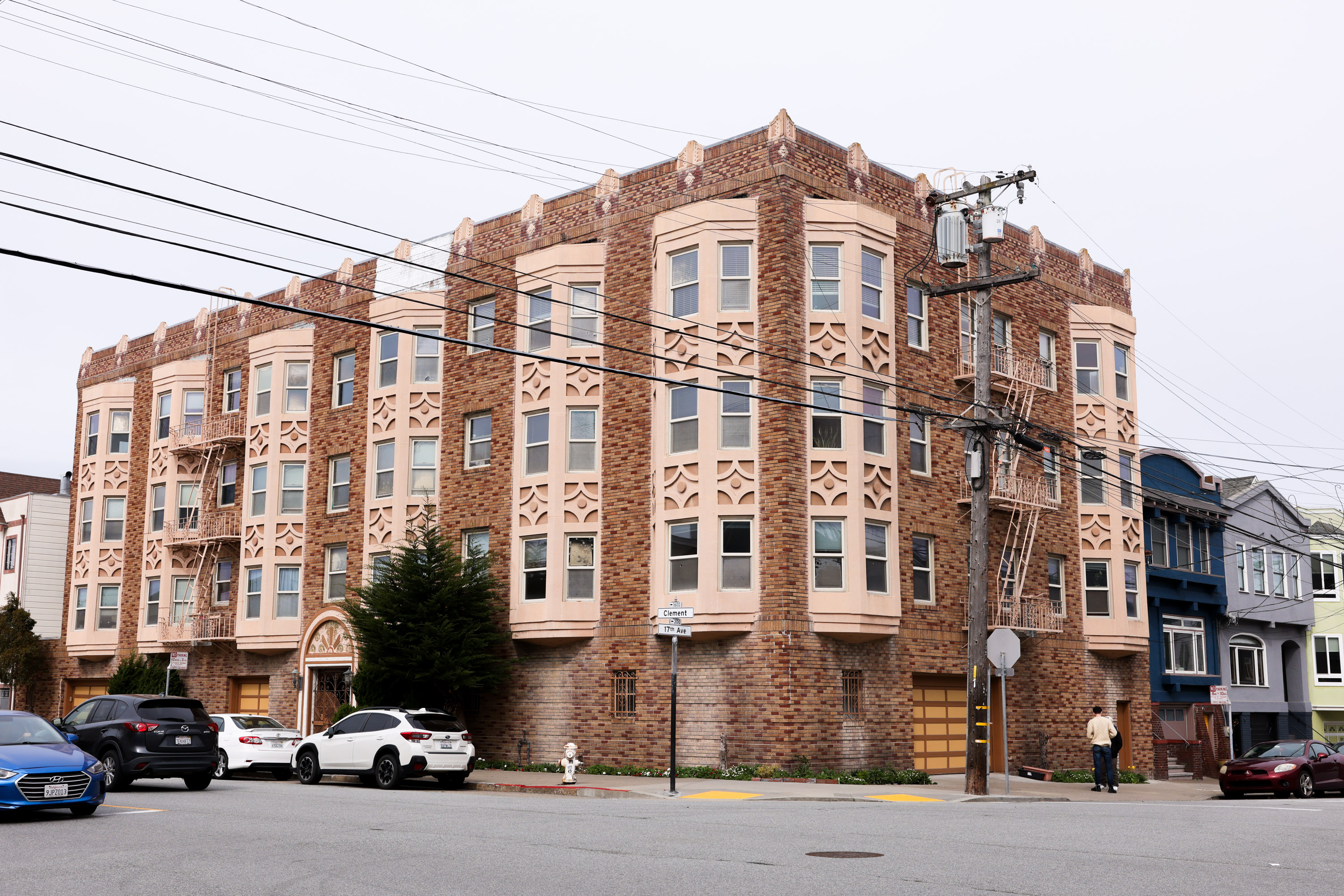 The image shows a brick apartment building with intricate designs around the windows, a fire escape, cars parked in front, and power lines crossing the scene.