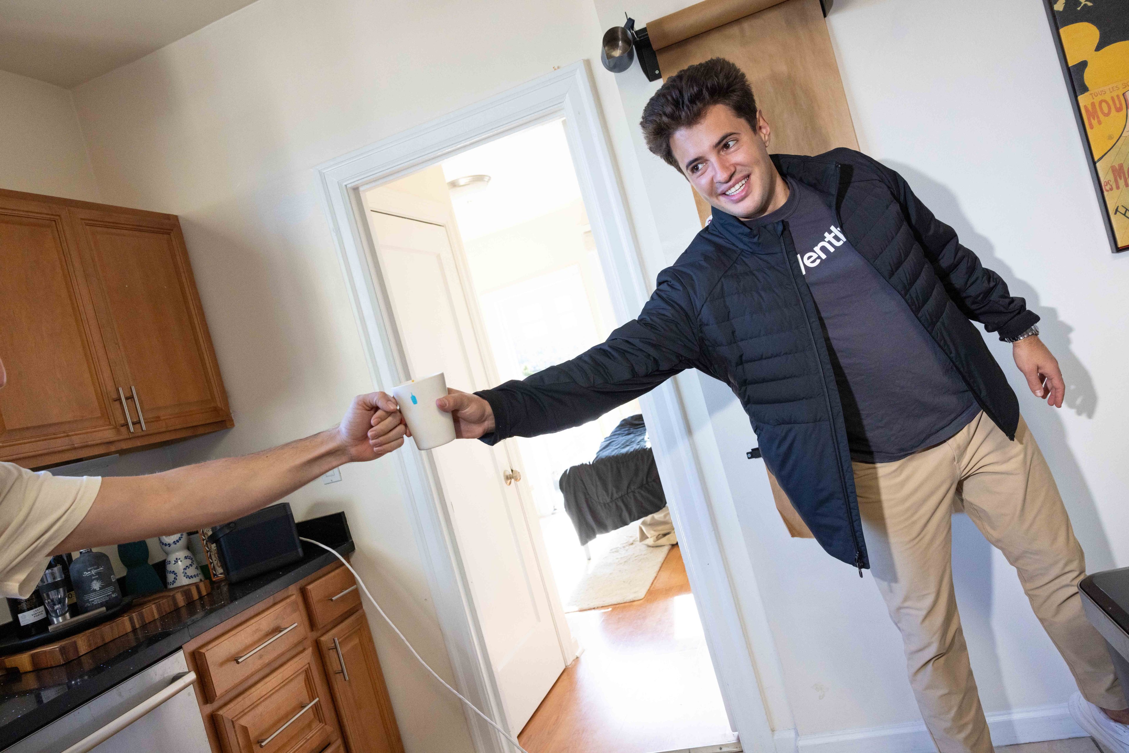 A smiling man in a black jacket and tan pants reaches from a room into a kitchen, exchanging a mug with another person. The kitchen has wooden cabinets.