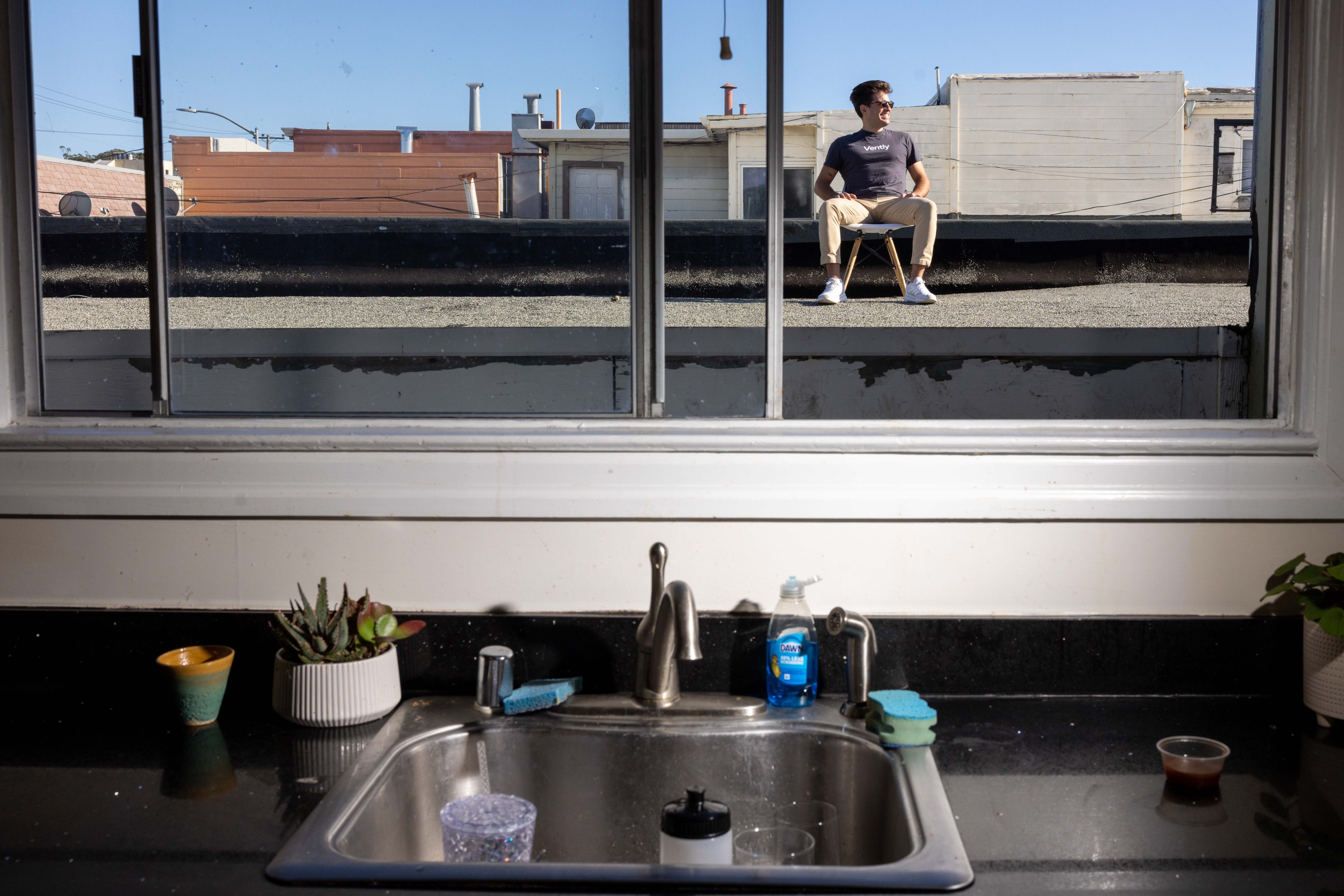 A man sits on a rooftop visible through a window above a kitchen sink, which has soap, sponges, and plants. The sky is clear and blue.