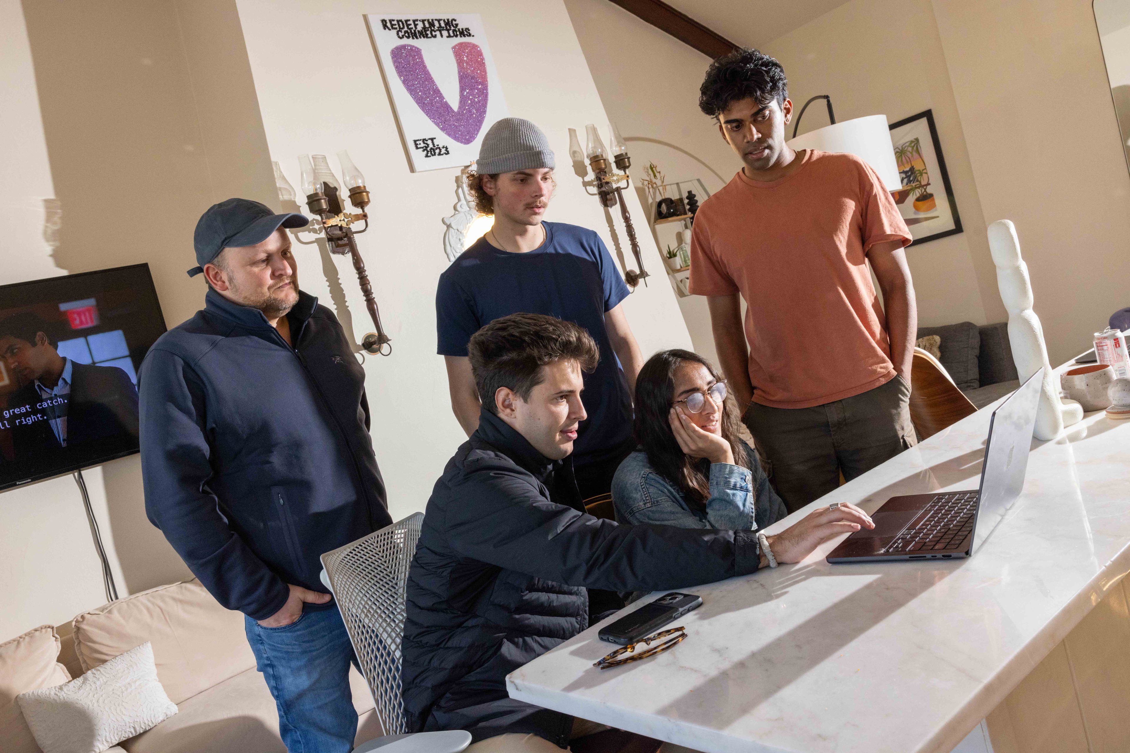 Five people gather around a table, looking intently at a laptop screen. Light streams in through a window, casting shadows on a marble countertop.