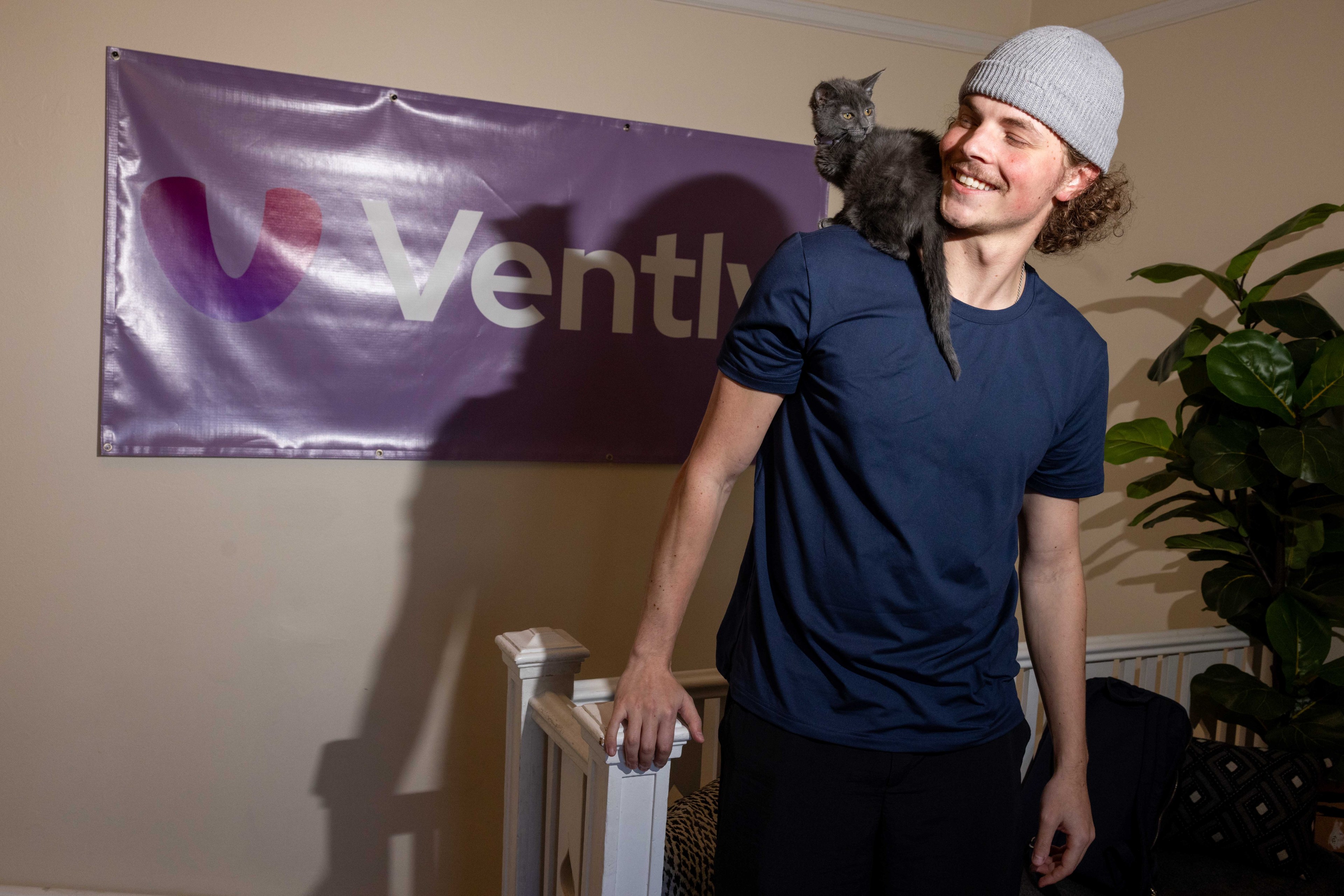 A smiling person in a gray beanie and navy shirt has a small black cat perched on their shoulder, standing next to a &quot;Vently&quot; banner and a plant.