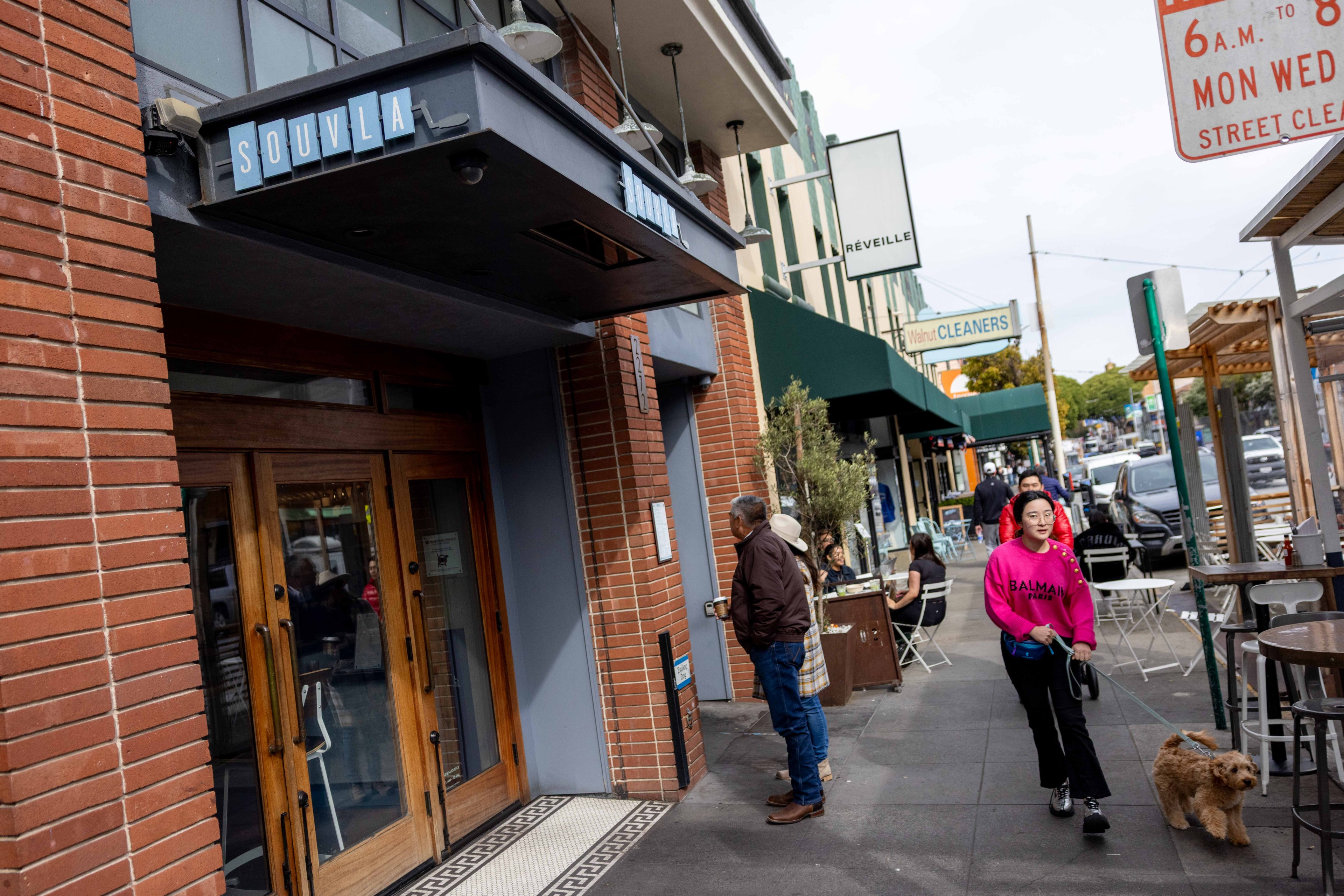 People stroll past a brick storefront labeled "Souvla." A woman in a pink hoodie walks a dog. The street has tables, greenery, and shops.