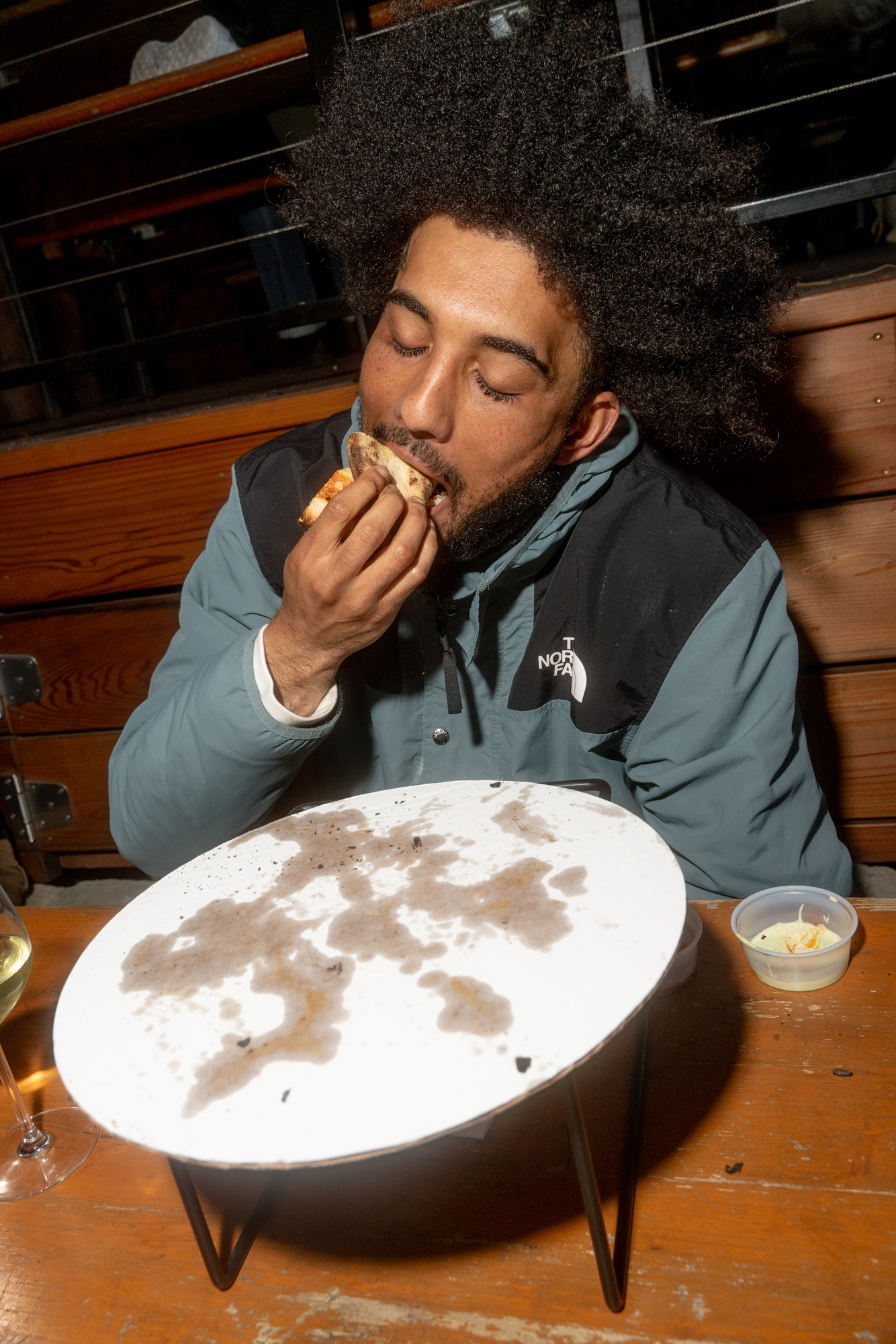 A person with curly hair is enjoying a slice of pizza. A dirty empty pizza tray, a drink, and a small container of coleslaw are on the table.