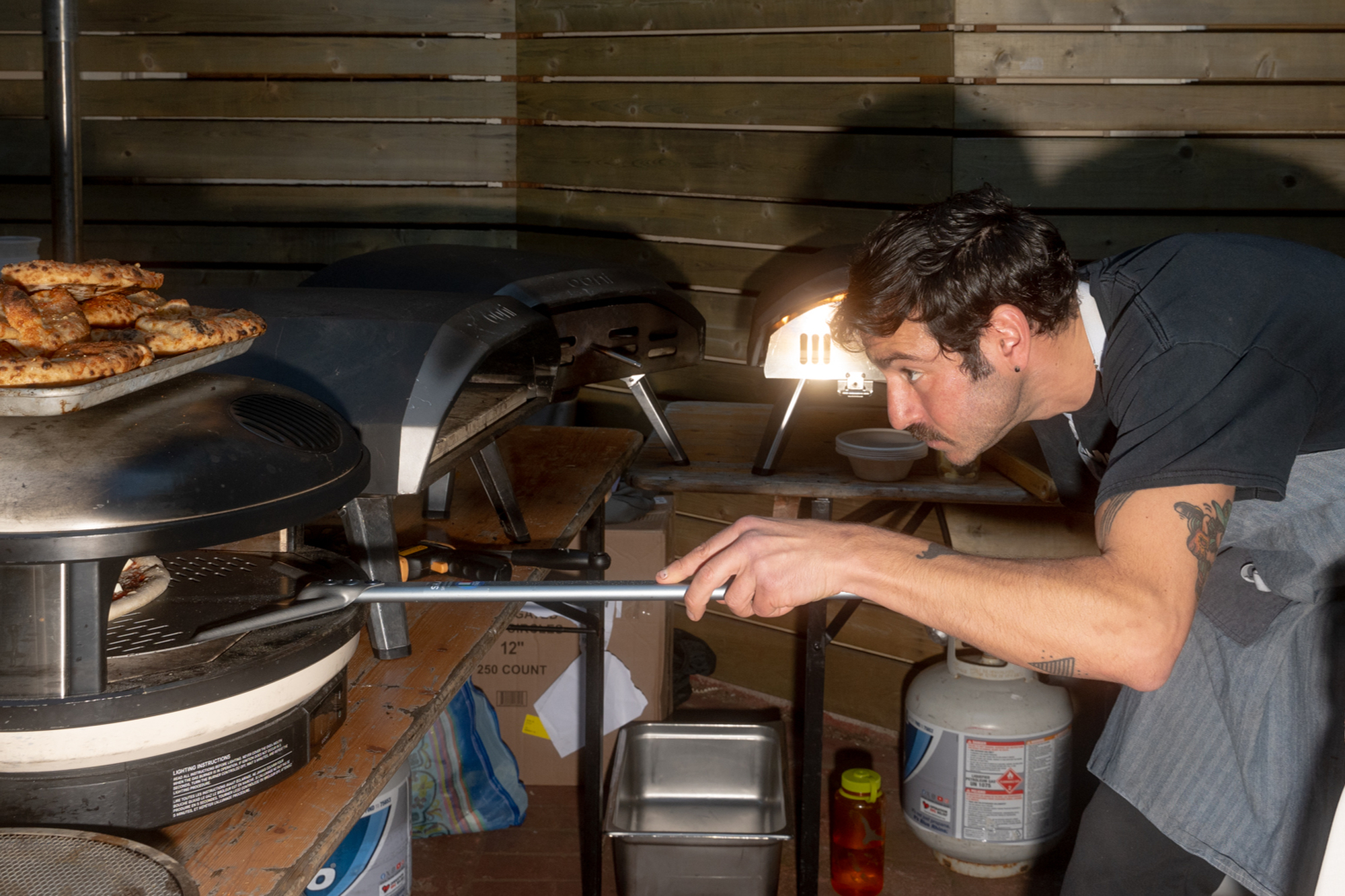 A person intently checks a pizza inside a wood-fired oven, holding a long peel. Nearby, a tray of pastries is on a counter in a rustic kitchen setting.