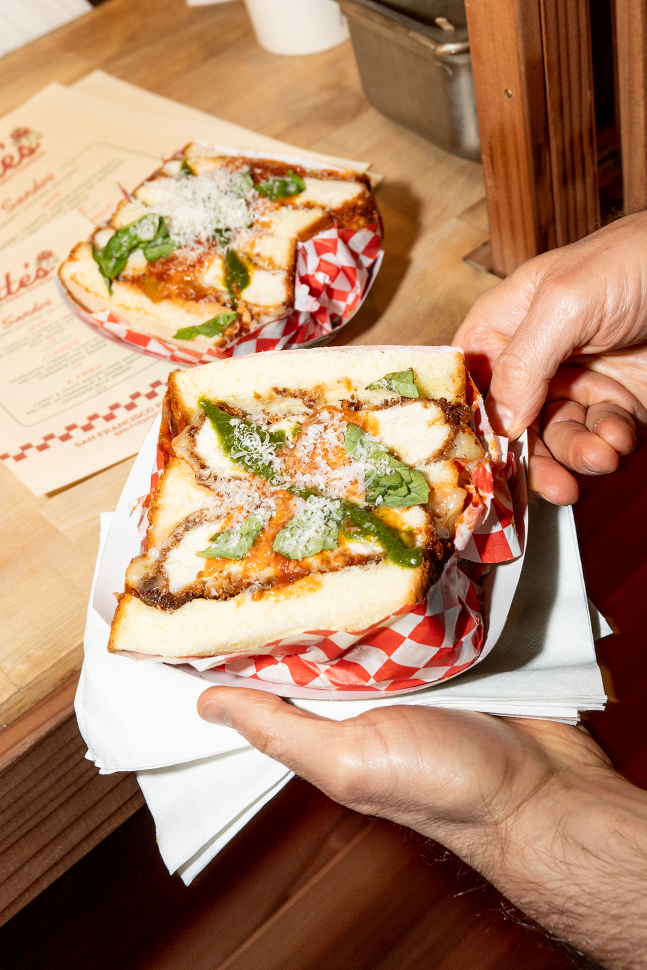 A person holds a sandwich with fried chicken, basil, tomato sauce, and cheese, served in checkered paper. Another similar sandwich is on the table.