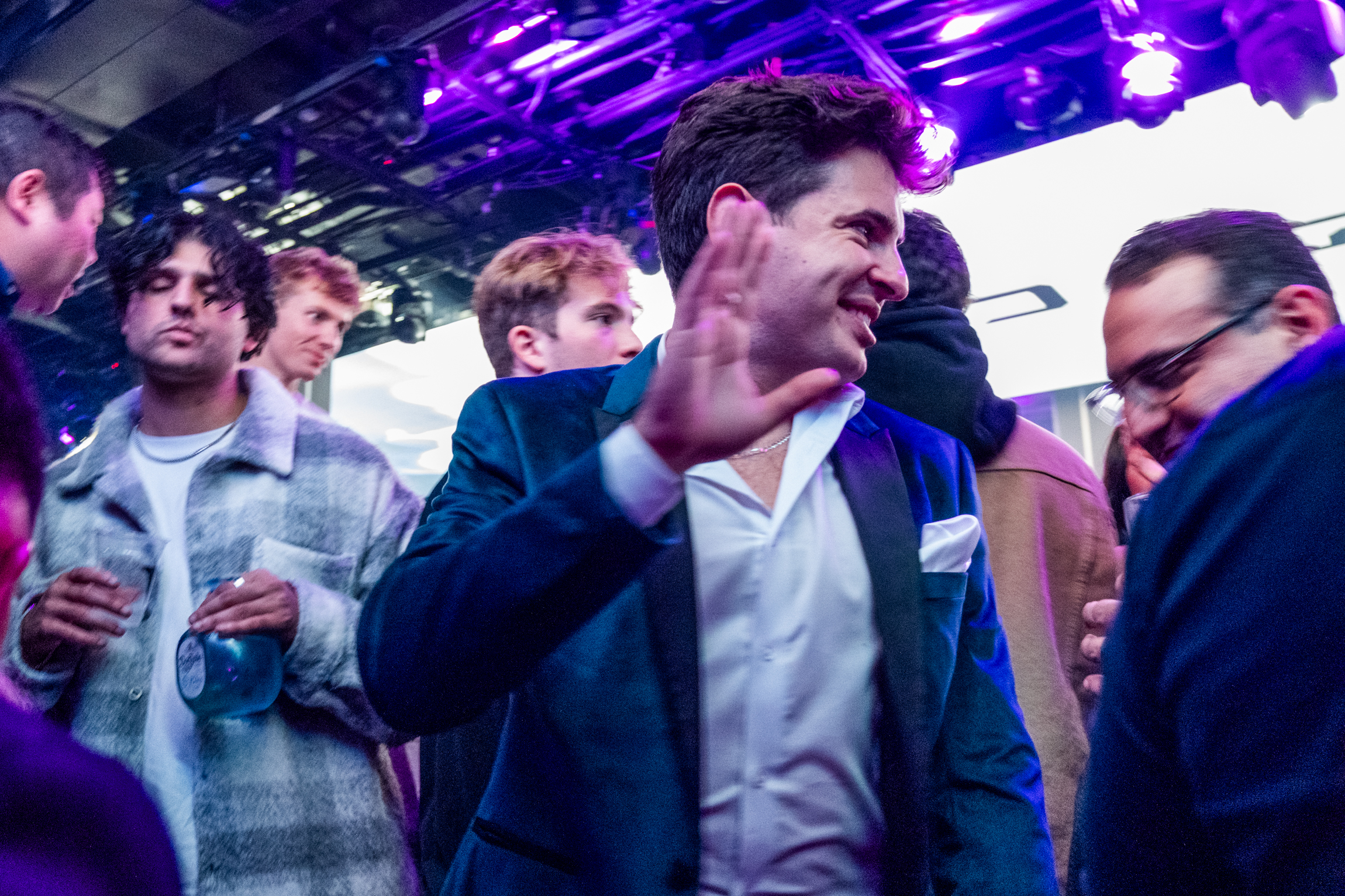 A group of people is enjoying a lively event under purple lights. One man in a dark blazer and white shirt is smiling and raising his hand in a high-five gesture.