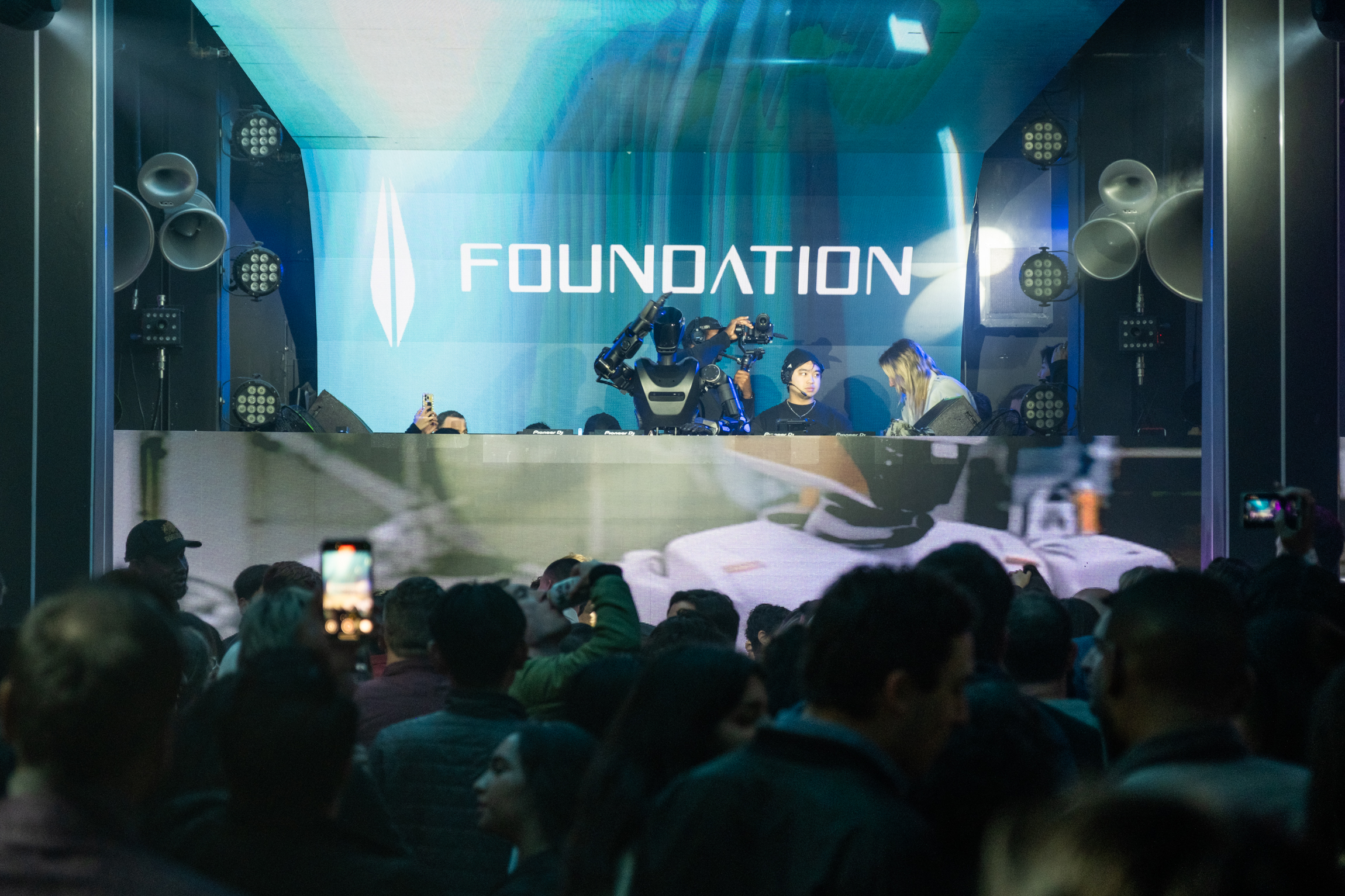 A crowd watches a robot DJ at a club event on stage under &quot;FOUNDATION&quot; signage. Bright lights and large speakers surround the lively atmosphere.