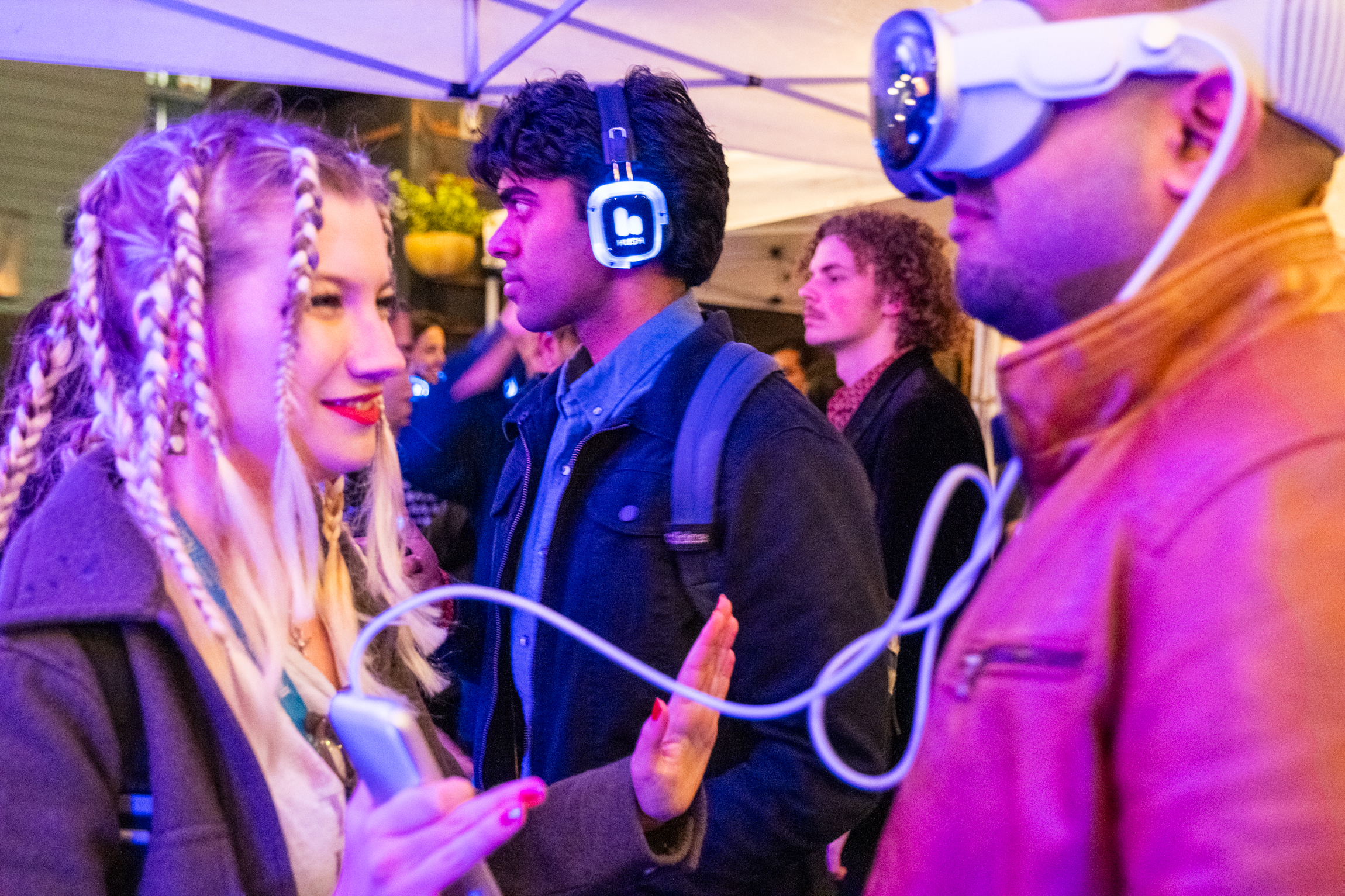 A lively scene with people wearing headphones, interacting. A woman with braided hair holds a device connected to a virtual reality headset worn by a man.