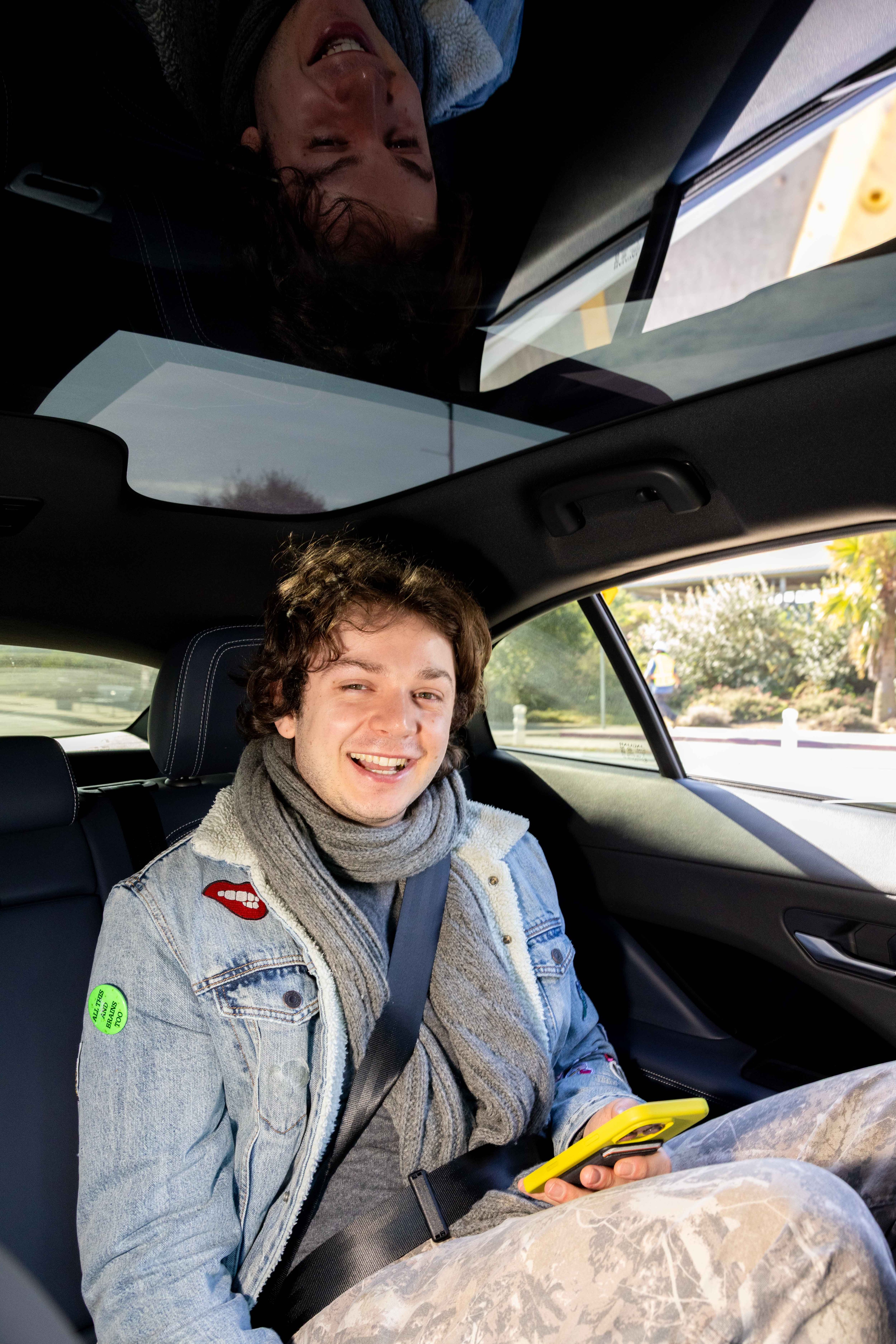 A person wearing a denim jacket, gray scarf, and holding a yellow phone smiles while seated in a car's rear seat. Sunlight highlights their casual attire.