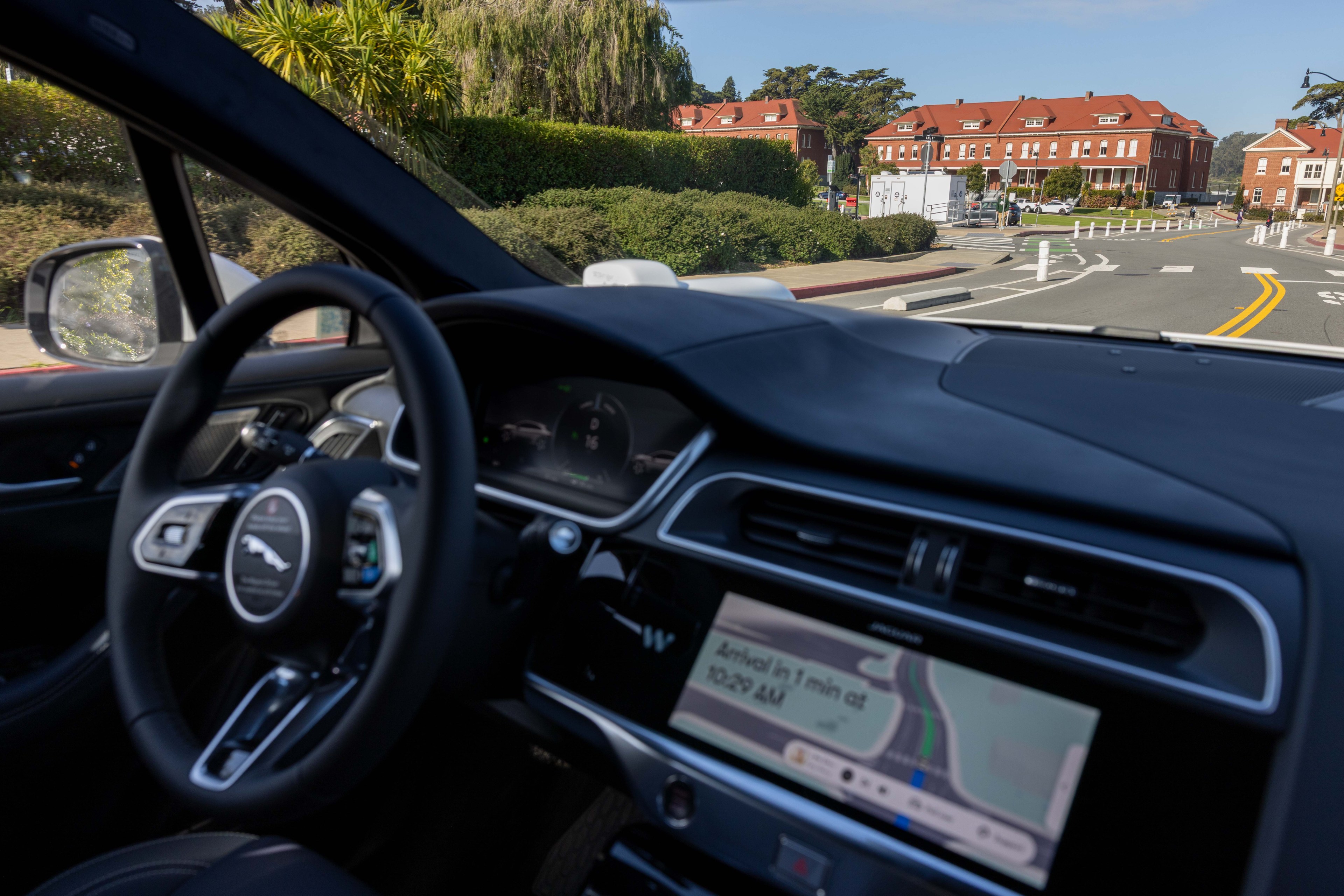 The image shows the interior of a car with a digital dashboard and GPS display, overlooking a suburban street lined with red-roofed brick buildings.