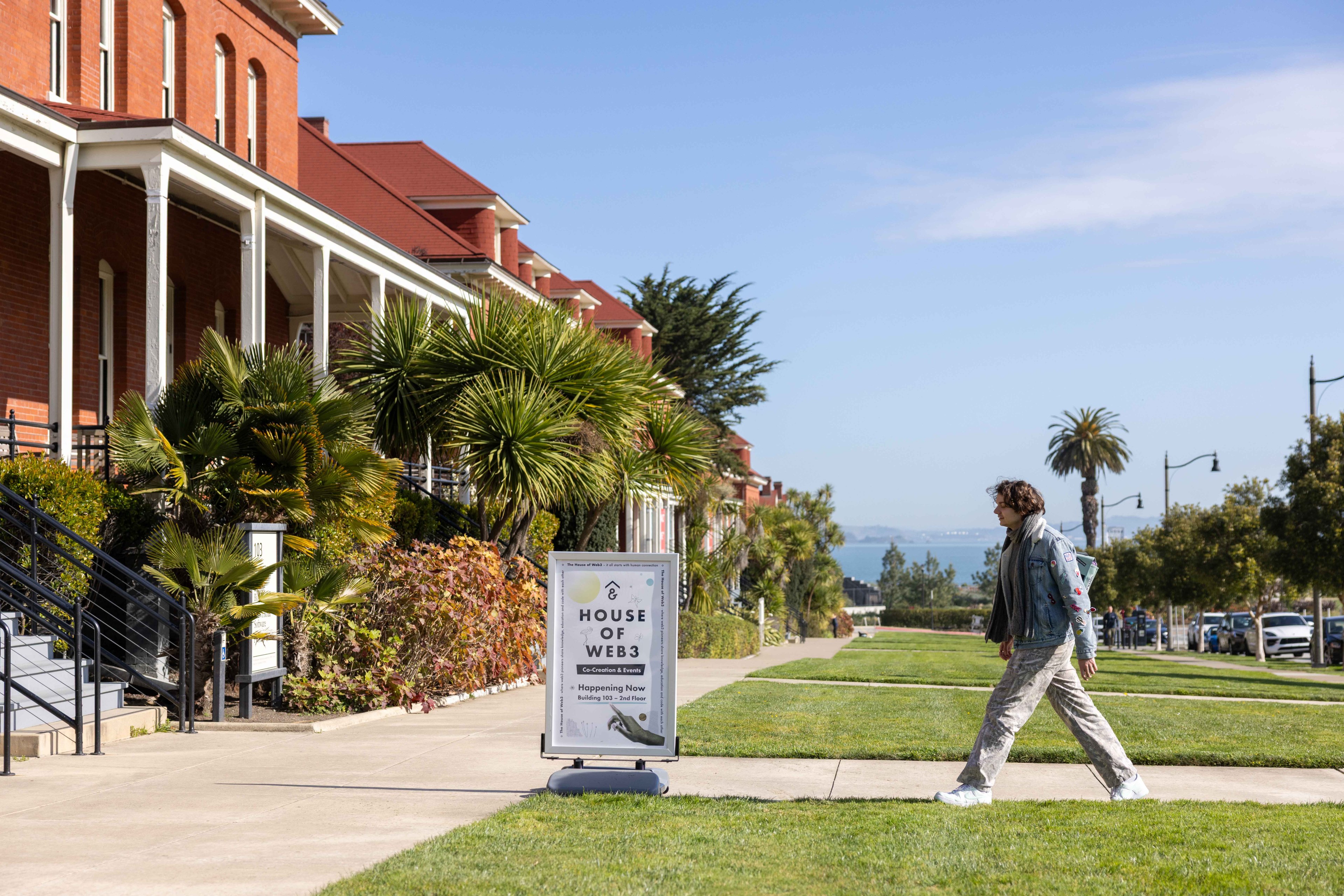 A person walks along a sidewalk lined with red brick buildings and lush greenery. A sign reads &quot;House of Web3.&quot; The sky is clear with distant water visible.