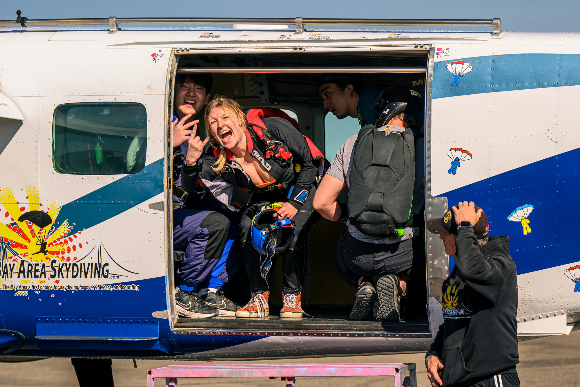 A skydiving group is in an airplane doorway. A smiling woman with pigtails gestures enthusiastically, while others prepare behind her. The plane has colorful graphics.