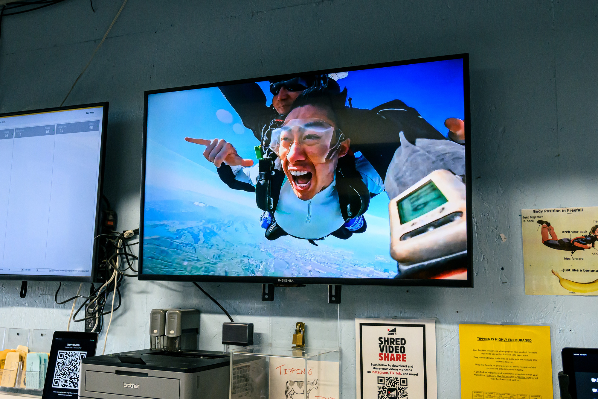 A TV screen displays a skydiving scene with a person excitedly free-falling tandem with an instructor, set against a blue sky and landscape background.