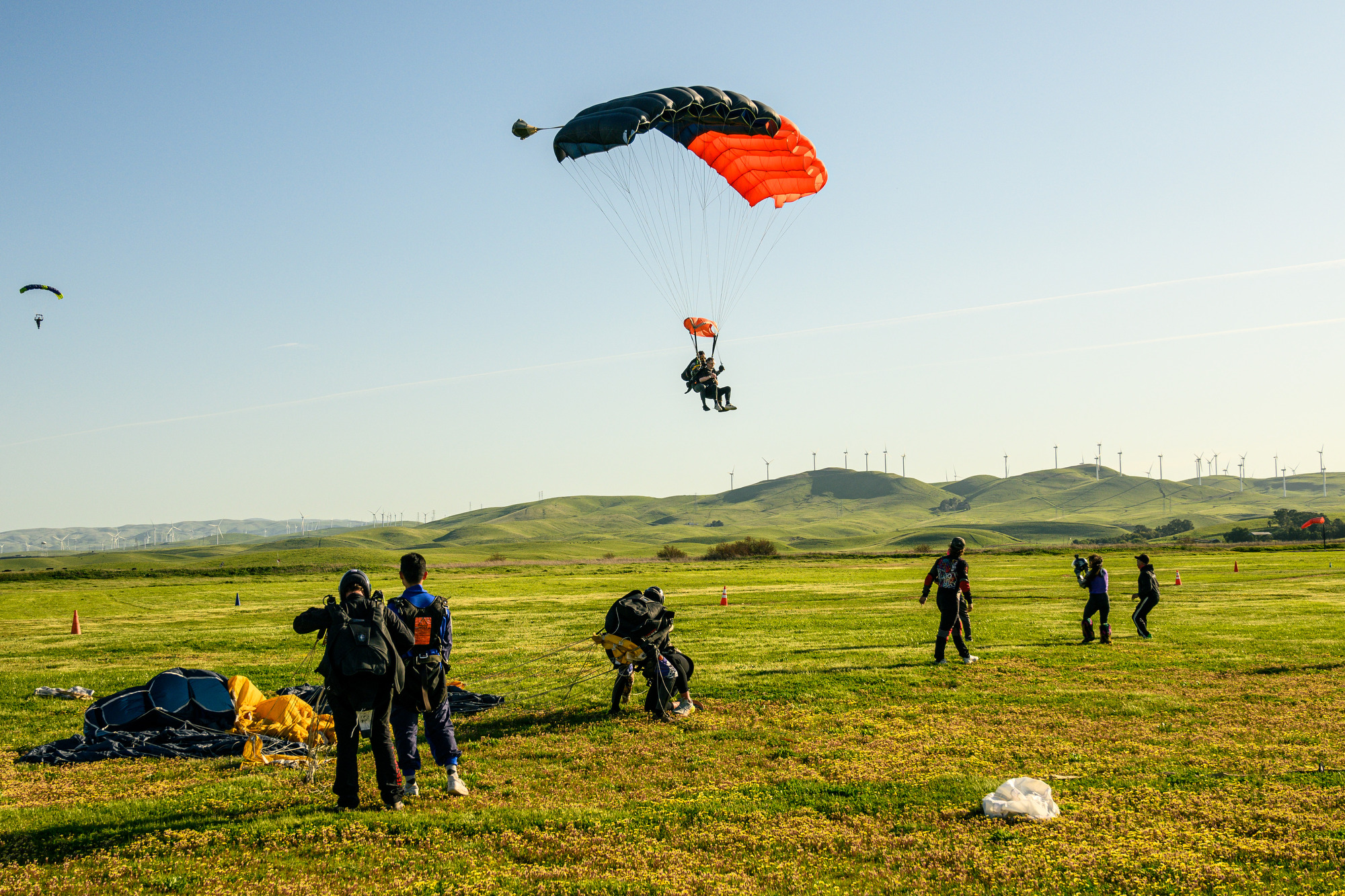 Skydivers land on a grassy field with parachutes open, while others prepare or watch, against a backdrop of green hills and wind turbines.