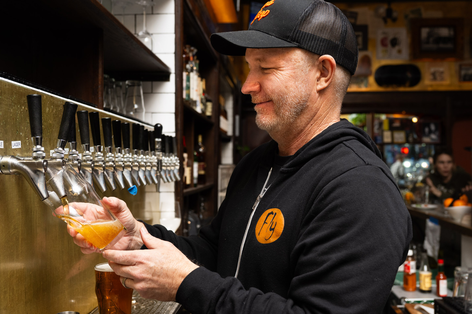A man in a black cap and jacket pours a pint of beer from a tap in a cozy, warmly lit bar with shelves of bottles and glasses behind him.